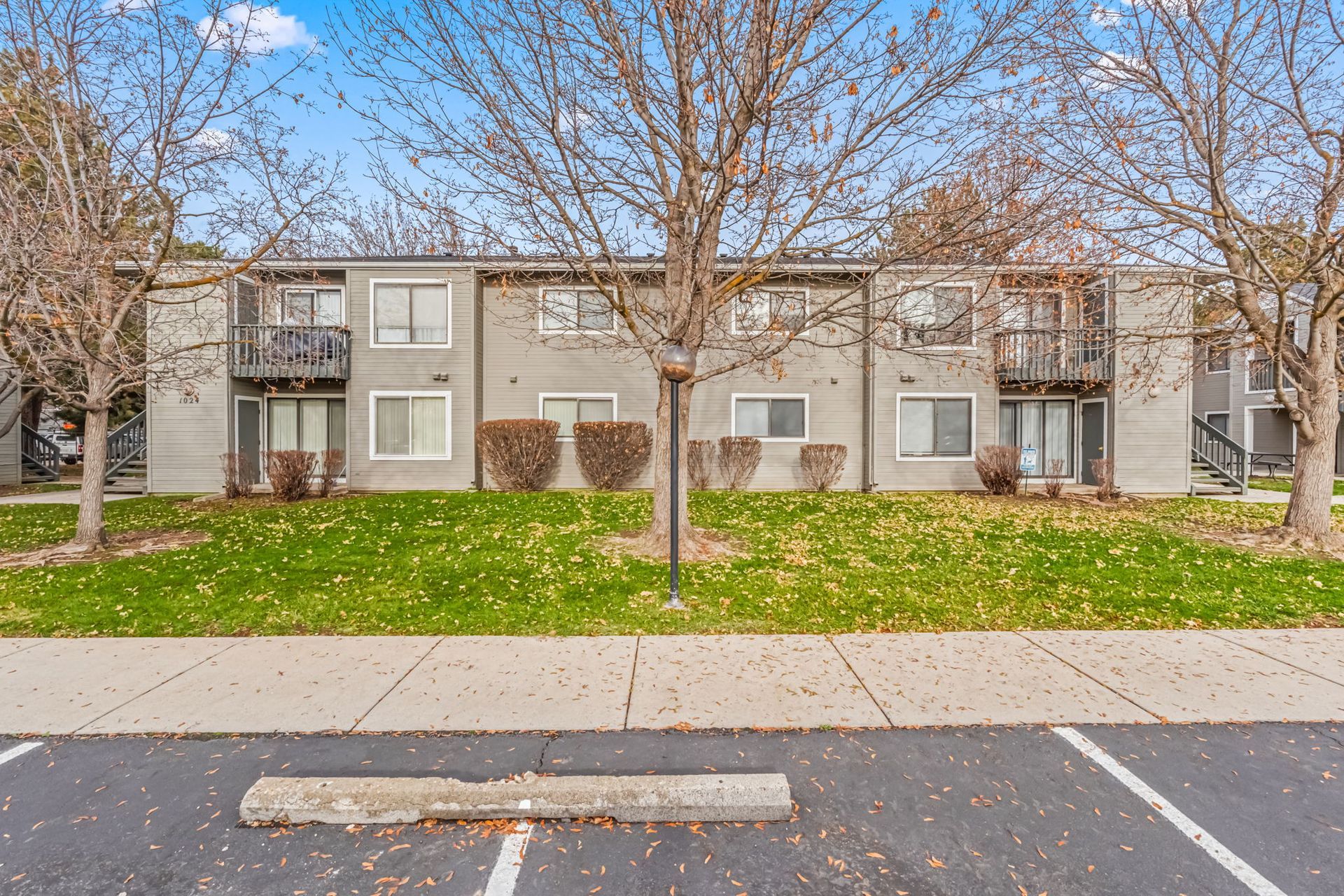 Gray two-story apartment building with balconies, trees, and a grassy lawn in front.