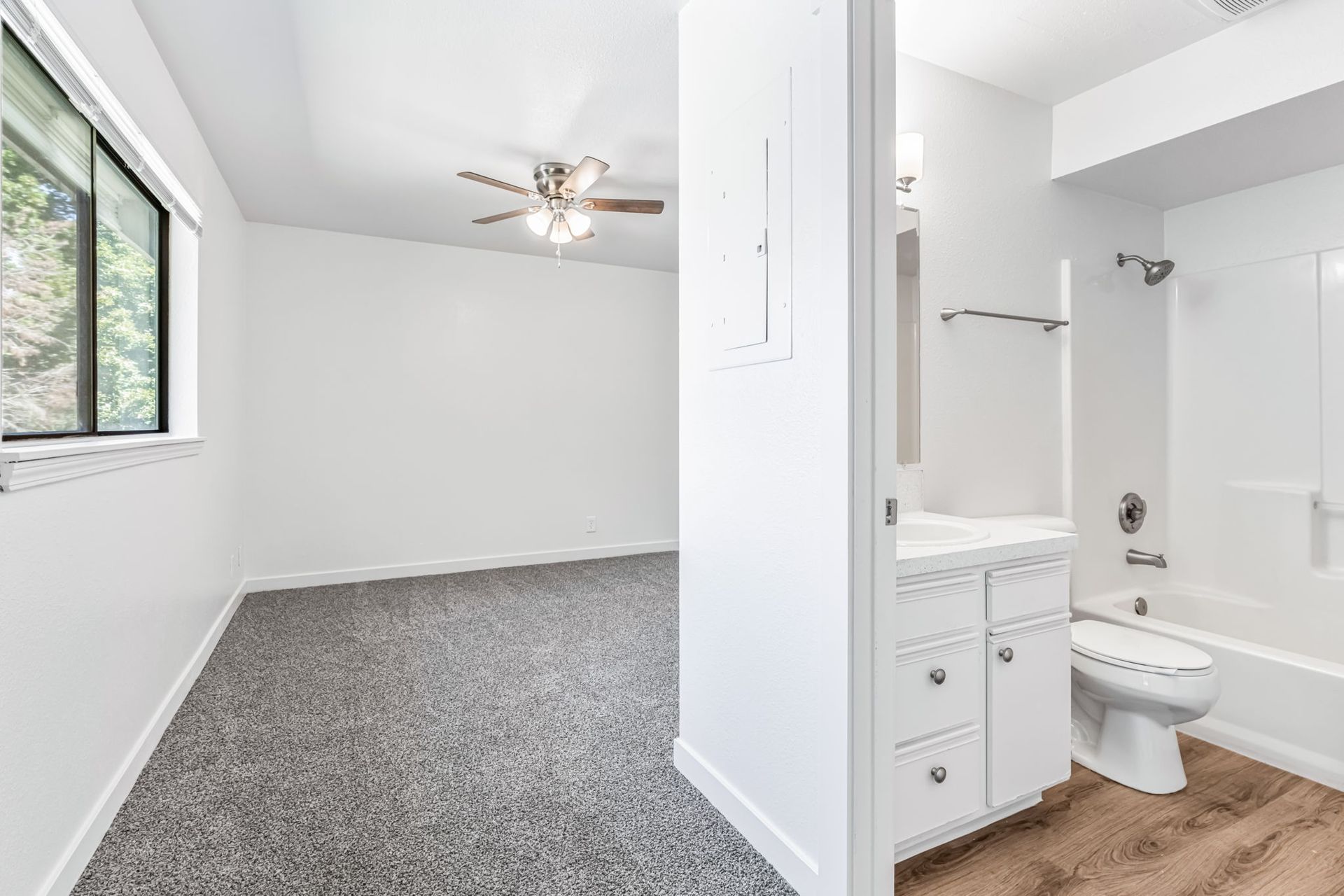 Empty, bright room with gray carpet, a bathroom visible with a toilet, sink, and tub.