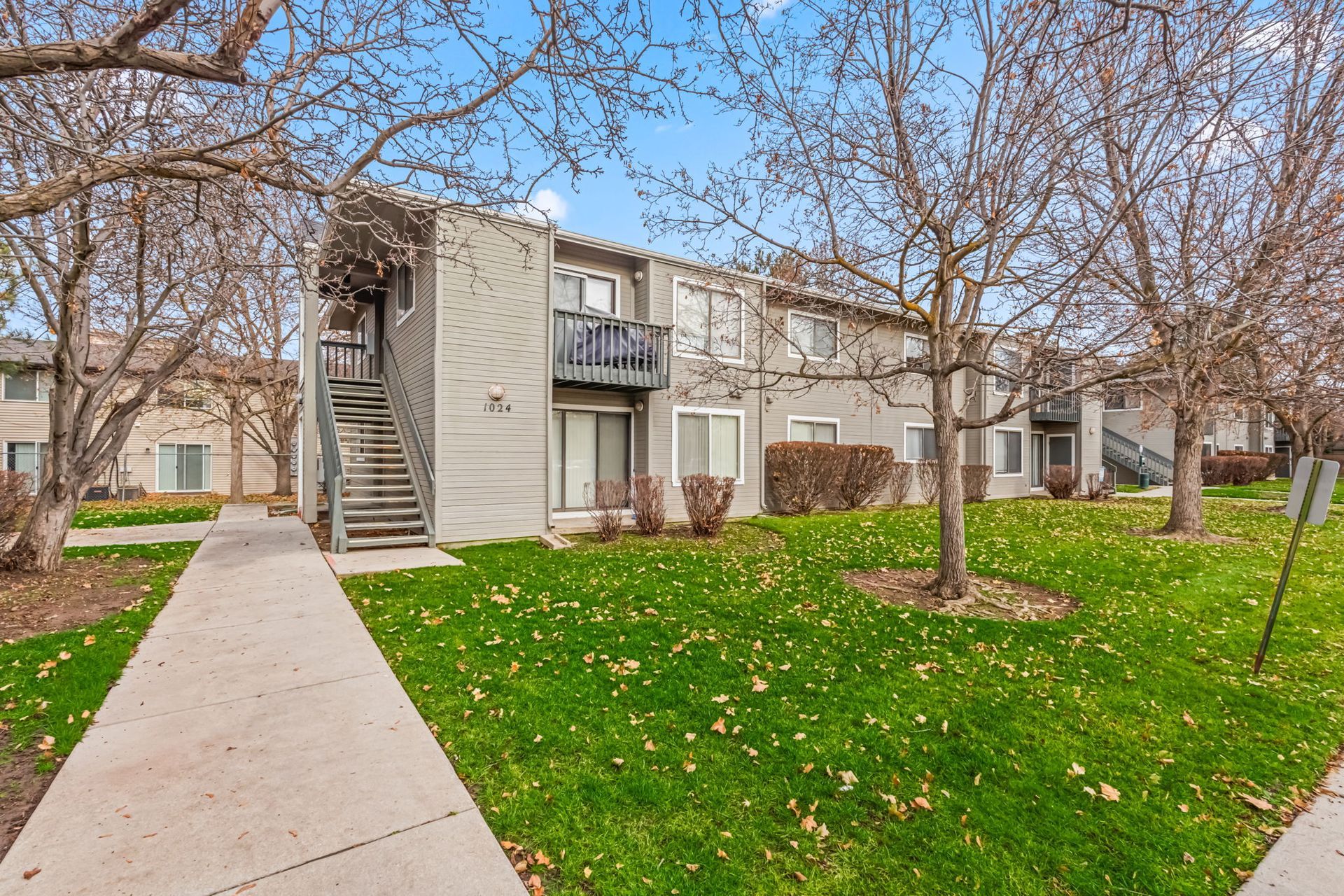 Two-story apartment building with stairwell and small balconies. Green lawn, bare trees, blue sky.