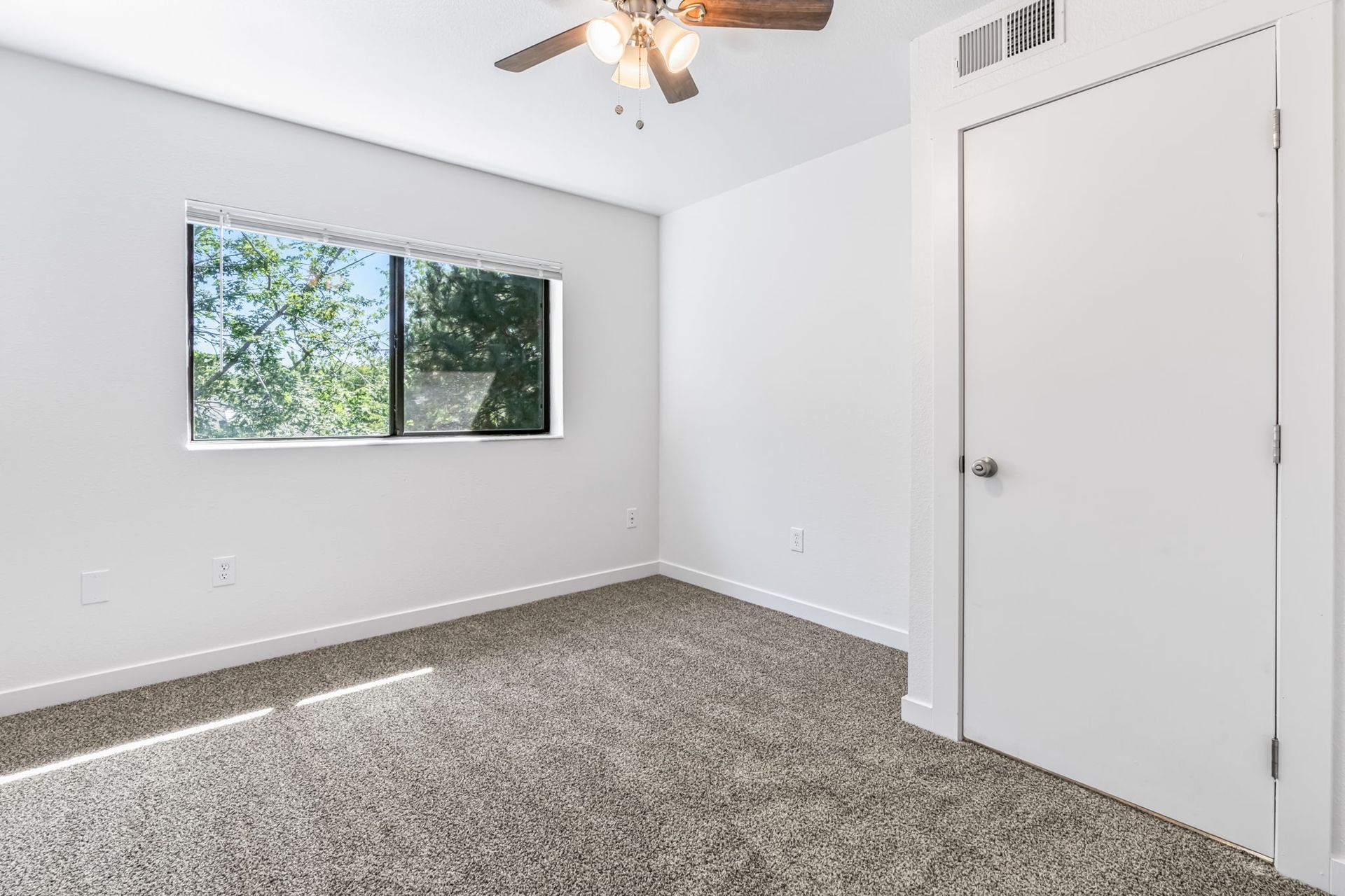 Empty bedroom with neutral carpet, white walls, window, door, and ceiling fan.