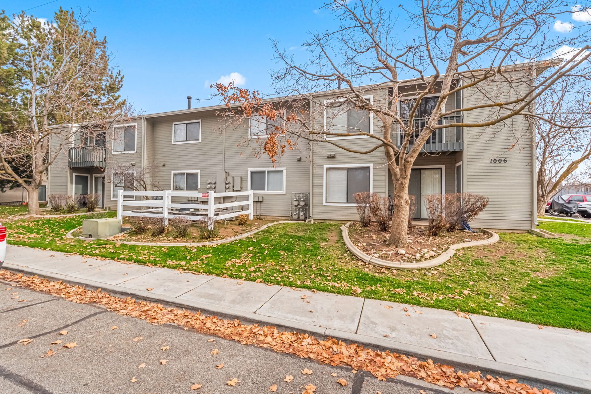 Two-story townhouses with gray siding, balconies, white fence, and fall foliage on a sunny day.