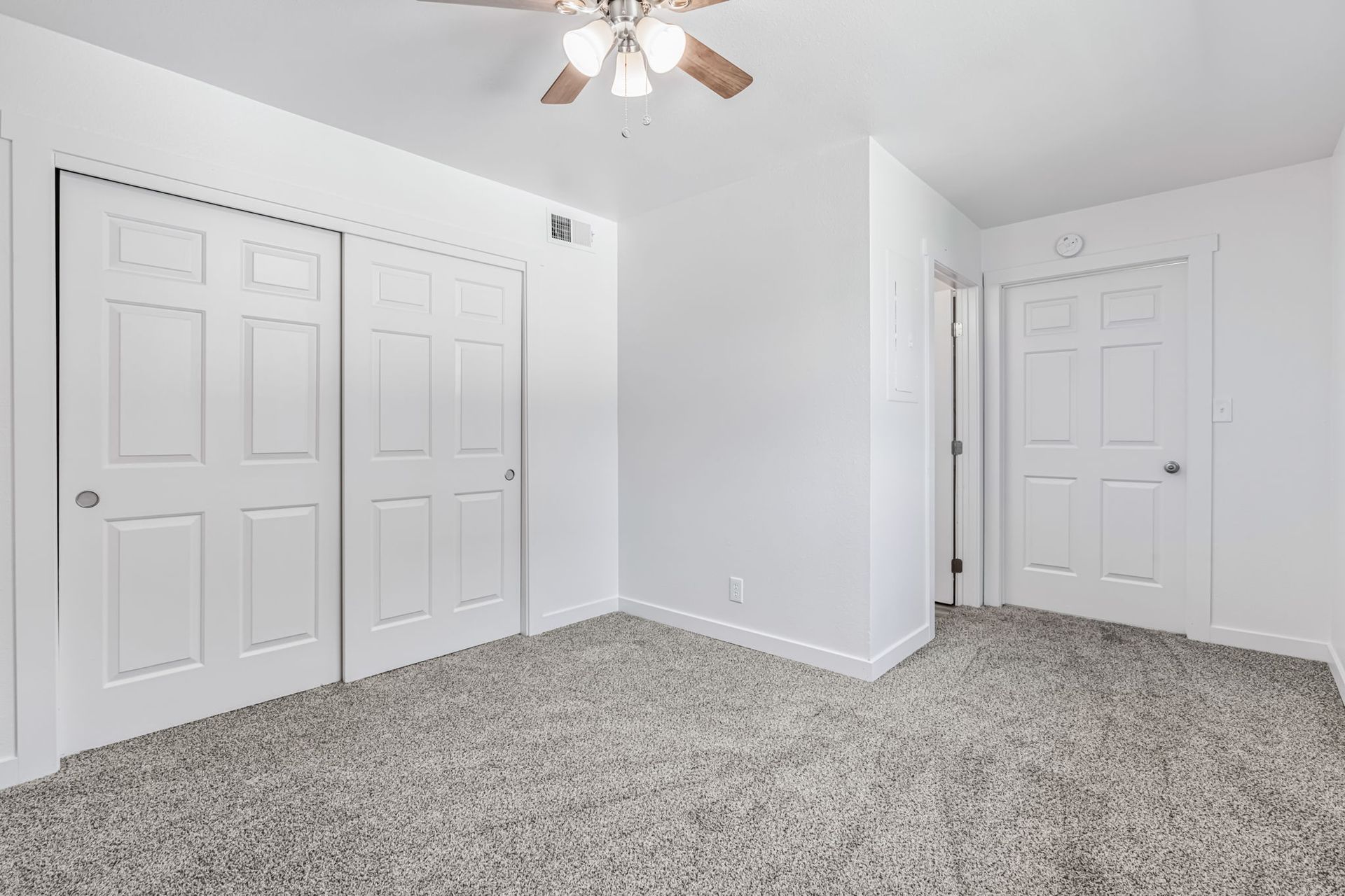 Empty bedroom with gray carpet, white walls, closet doors, and a ceiling fan.