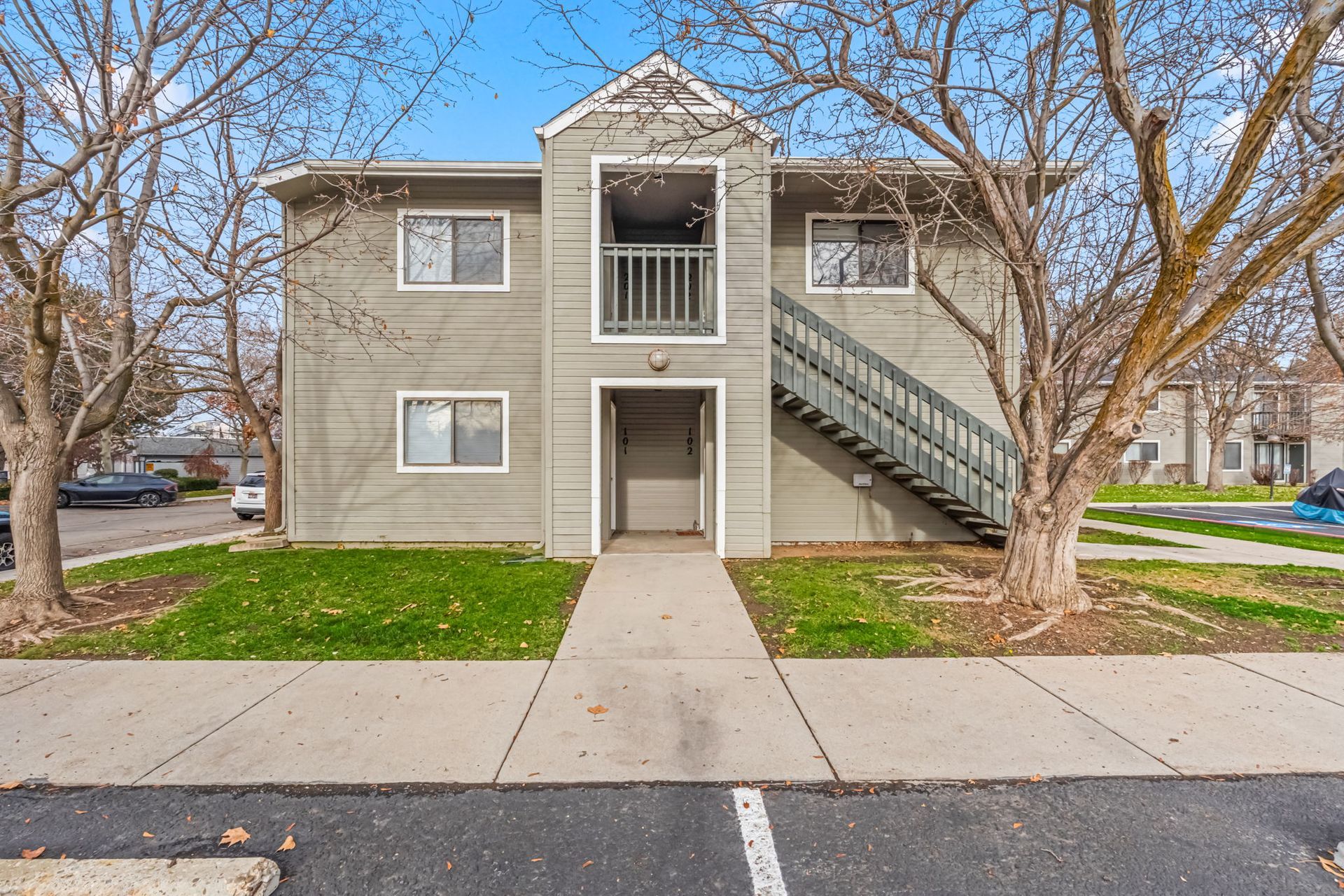 Two-story apartment building with stairs and a balcony. Gray siding, pathway to the front door, trees in the yard.
