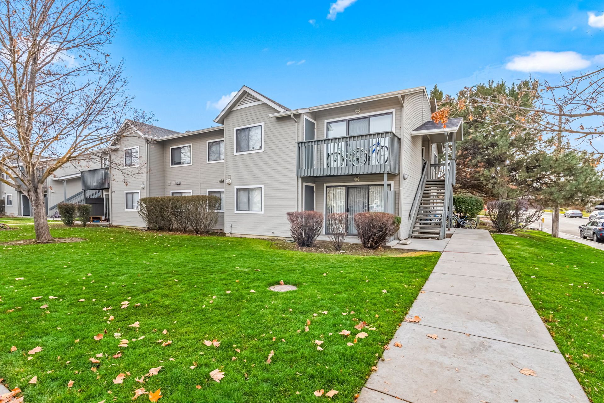 Two-story apartment building with balconies, gray siding, green lawn, sidewalk, and blue sky.