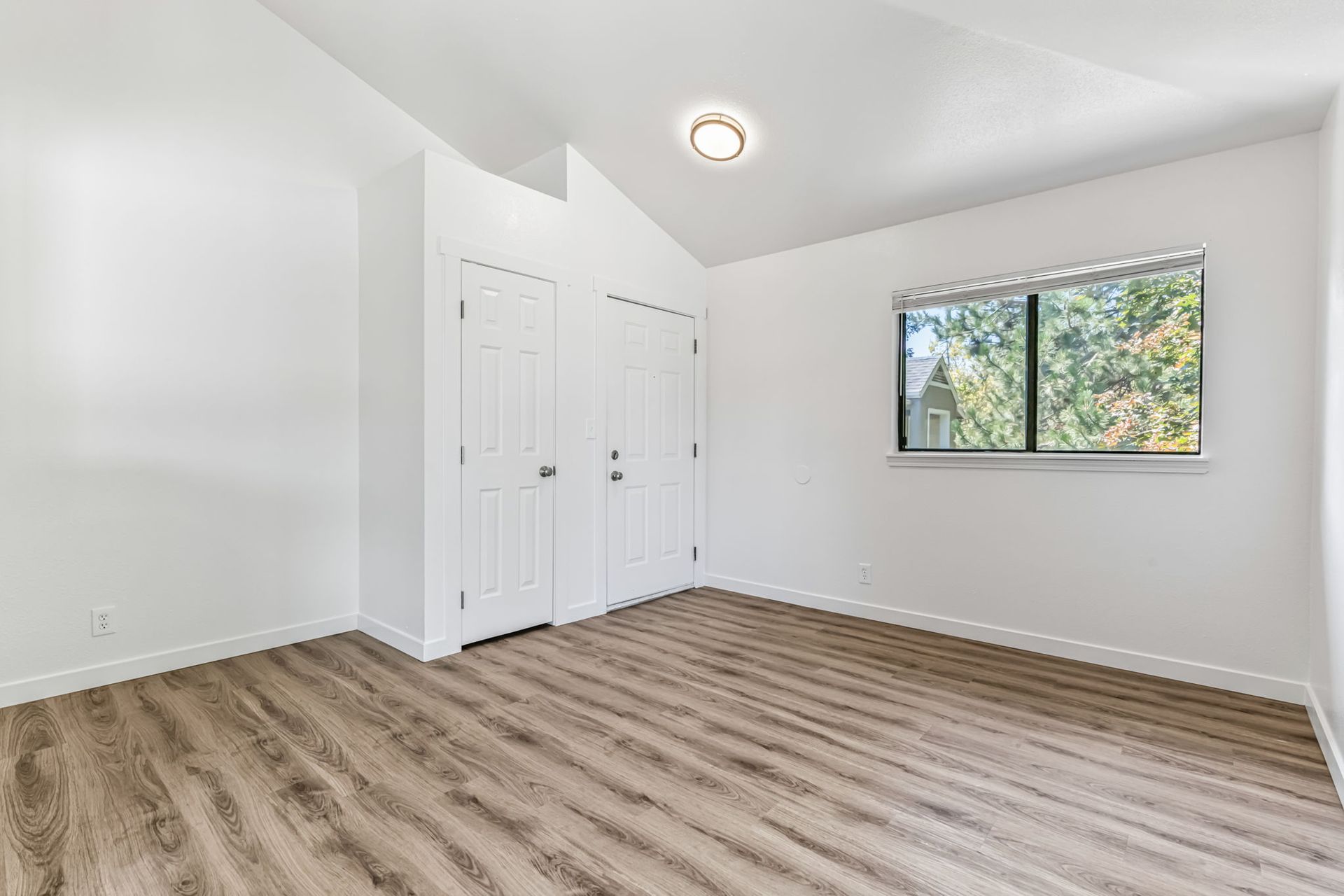 Empty room with white walls, two closet doors, window with trees outside, and wood-look flooring.