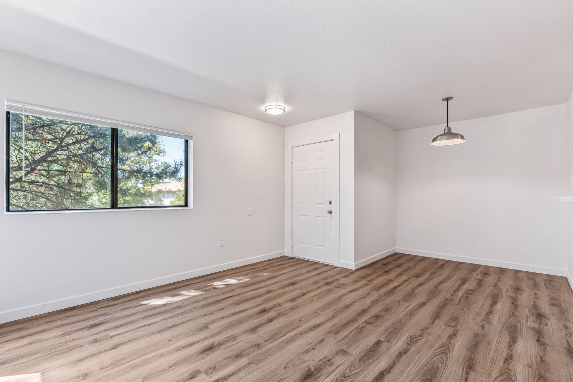 Empty room with wood-look flooring, white walls, window with view of trees, and two light fixtures.