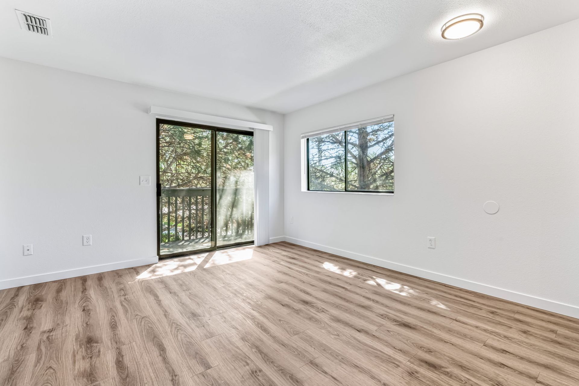 Empty room with light wood floor, sliding glass door to a balcony, and window with outdoor view.