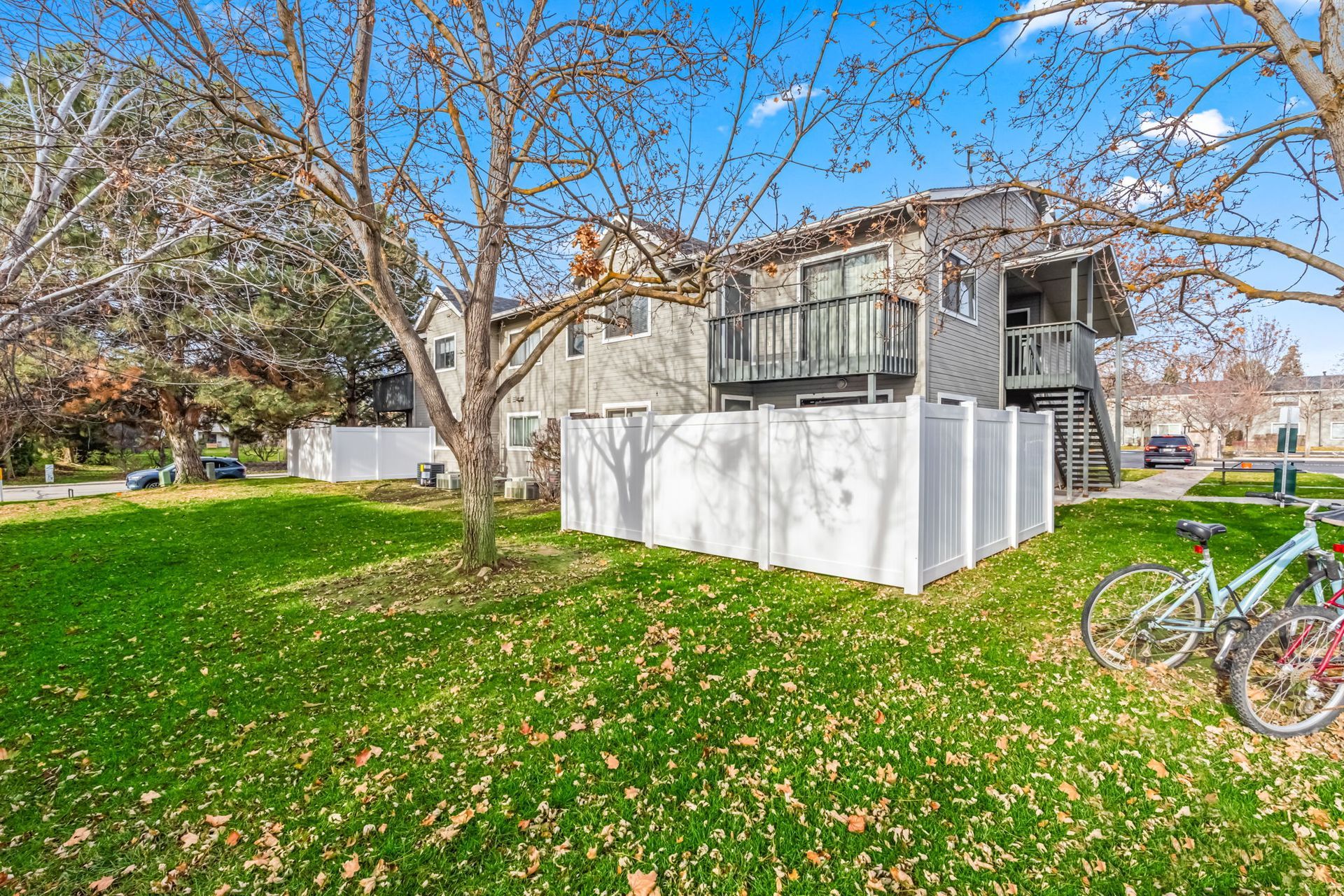 Two-story apartment building with balcony, white fence, and two bikes on green lawn under a blue sky.