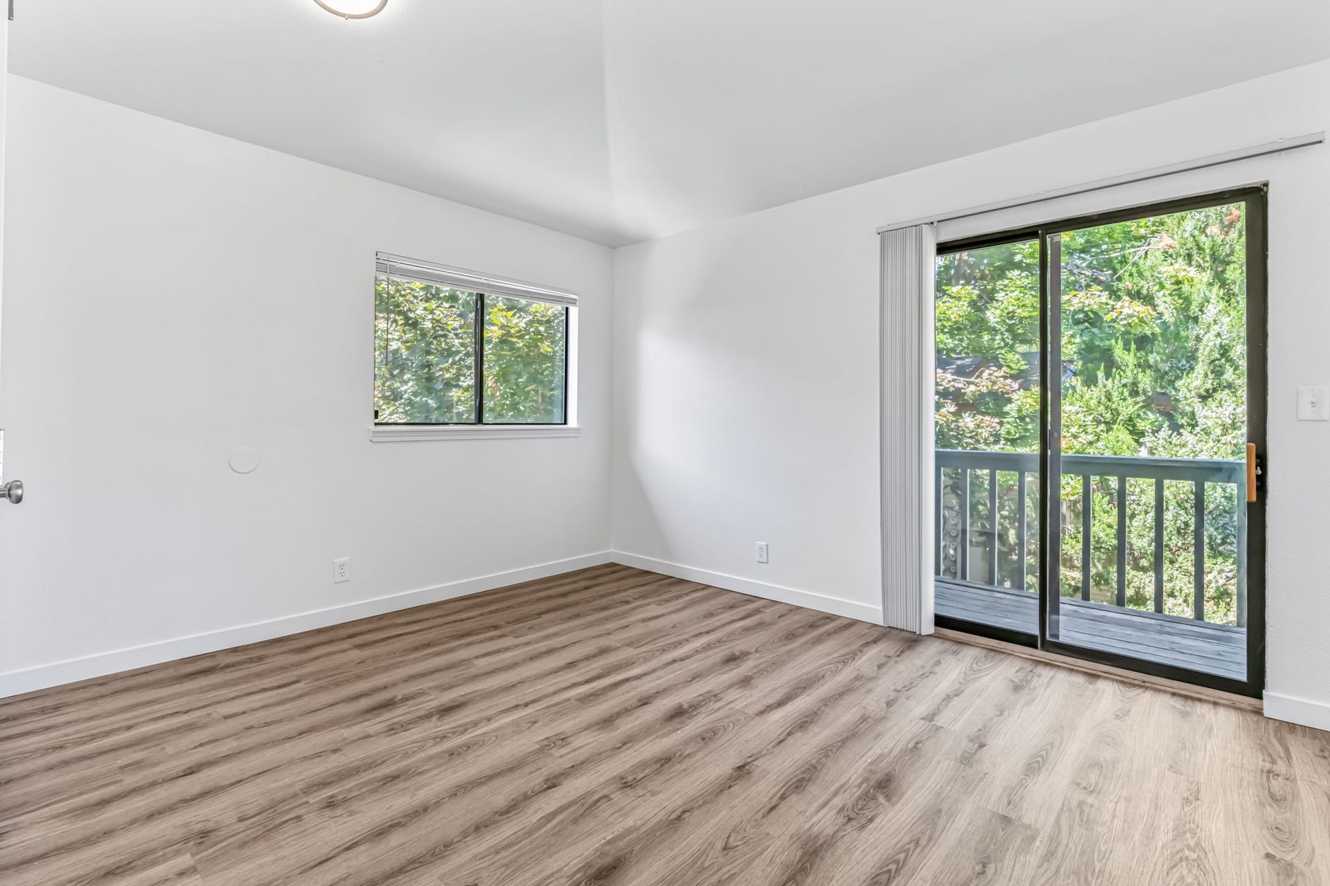 Empty bedroom with wooden floor, sliding glass door to balcony with trees, and small window.