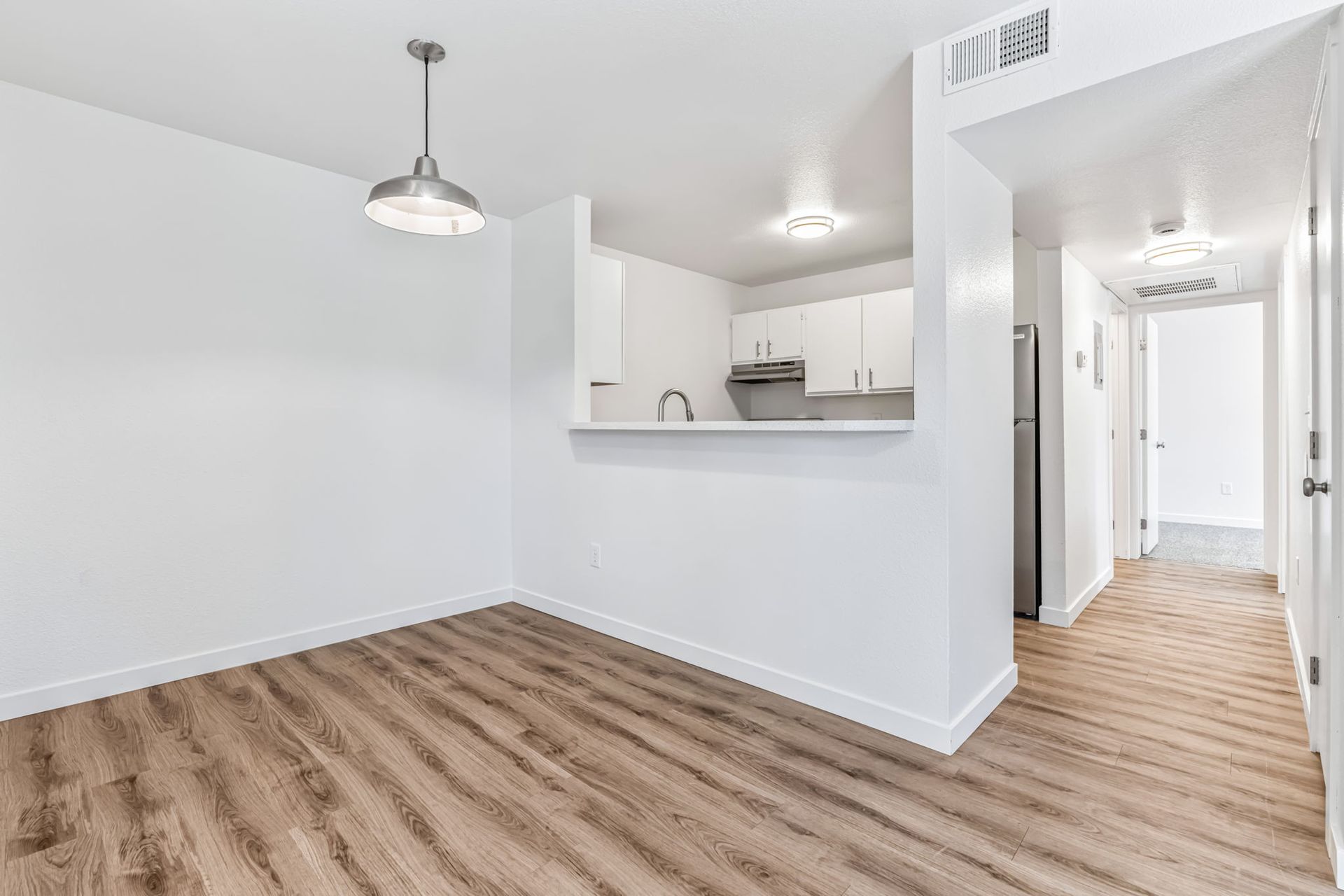 Empty, white-walled room with wooden floor and a small kitchen visible in the background; a hanging light.