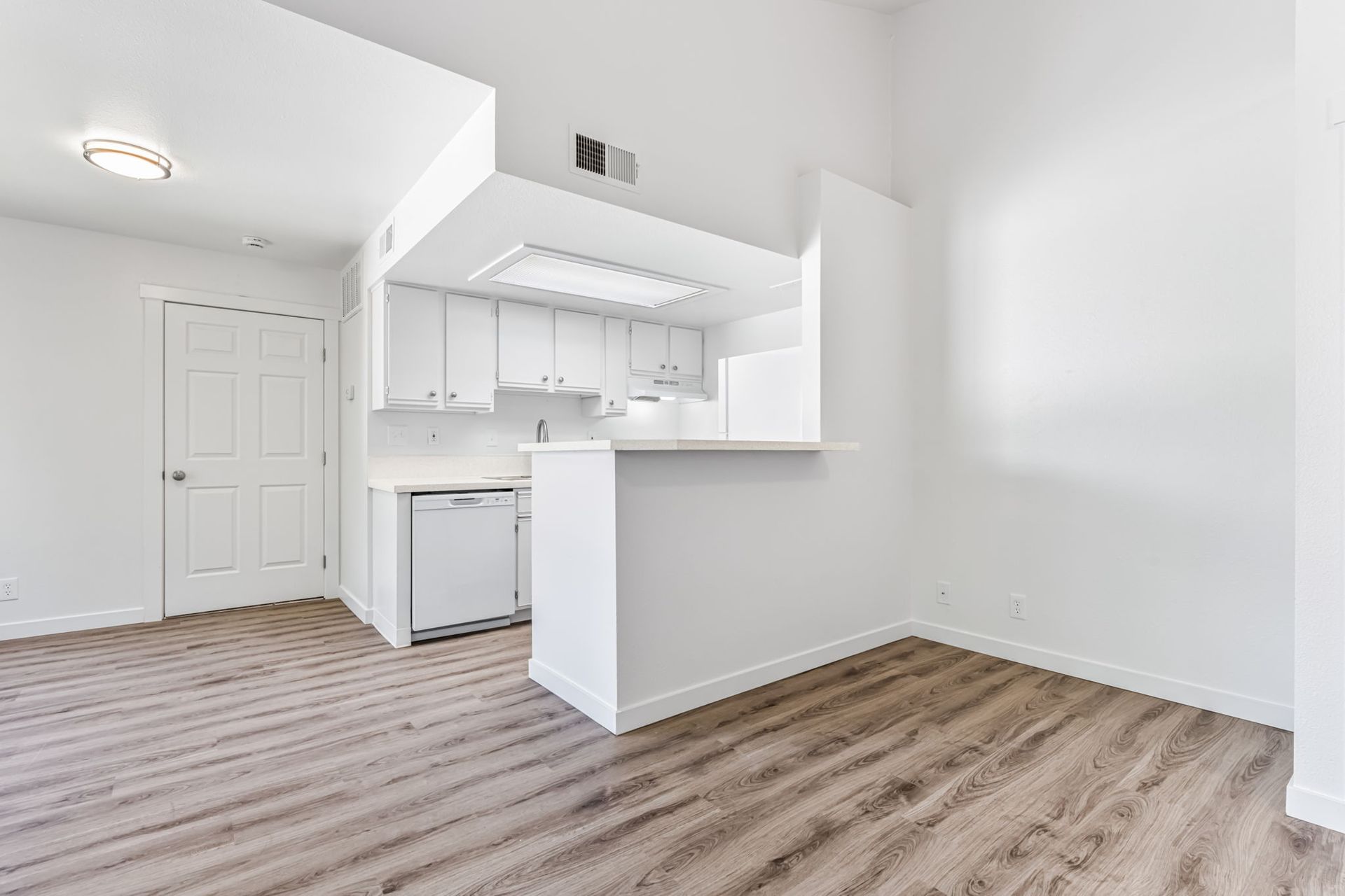 Empty, white-walled apartment with a small kitchen and wooden-look flooring.