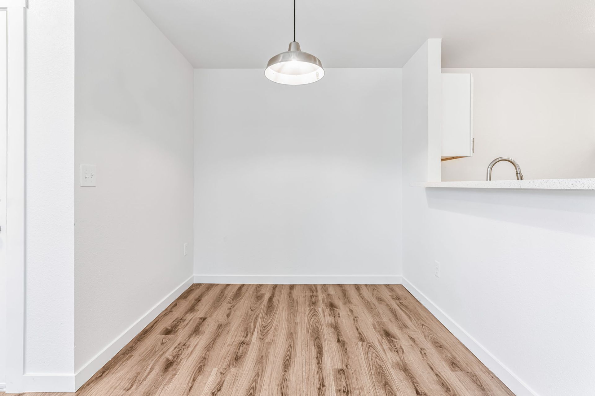 Empty white-walled dining area with wood-look flooring and a pendant light. Partial kitchen visible.