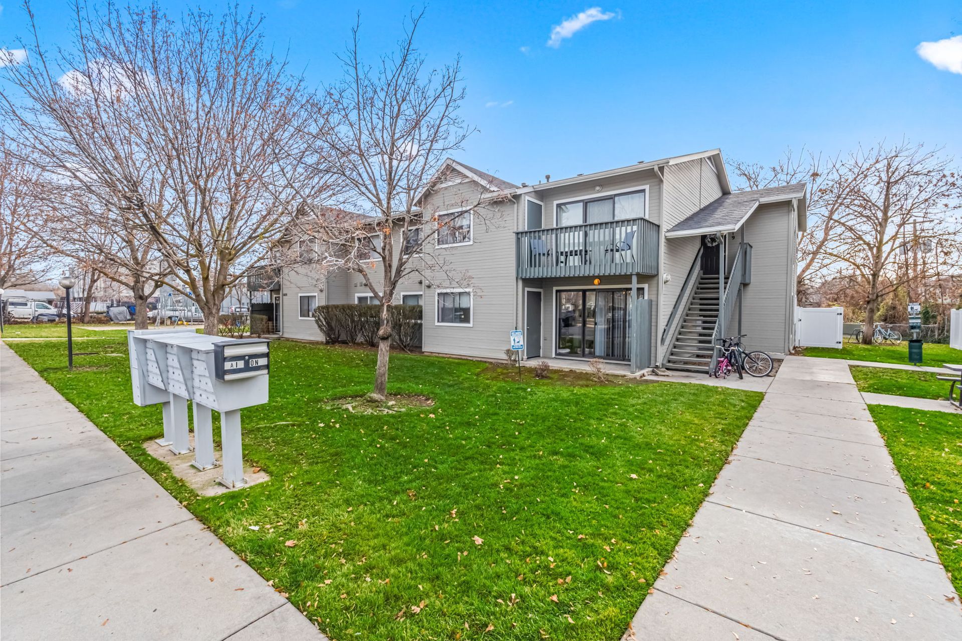 Apartment building exterior, two-story, gray siding, green lawn, mailbox, sidewalk, and trees under a blue sky.