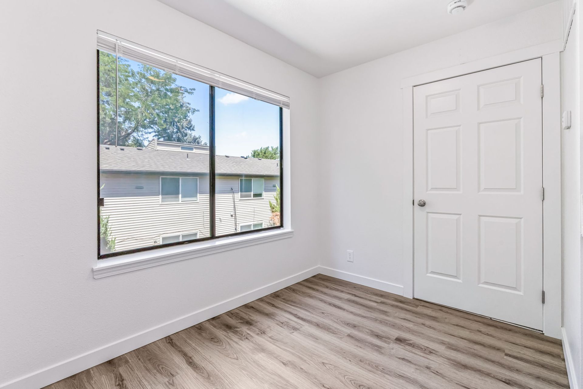 Empty white room with a window overlooking houses and a closed door. Wooden floor.