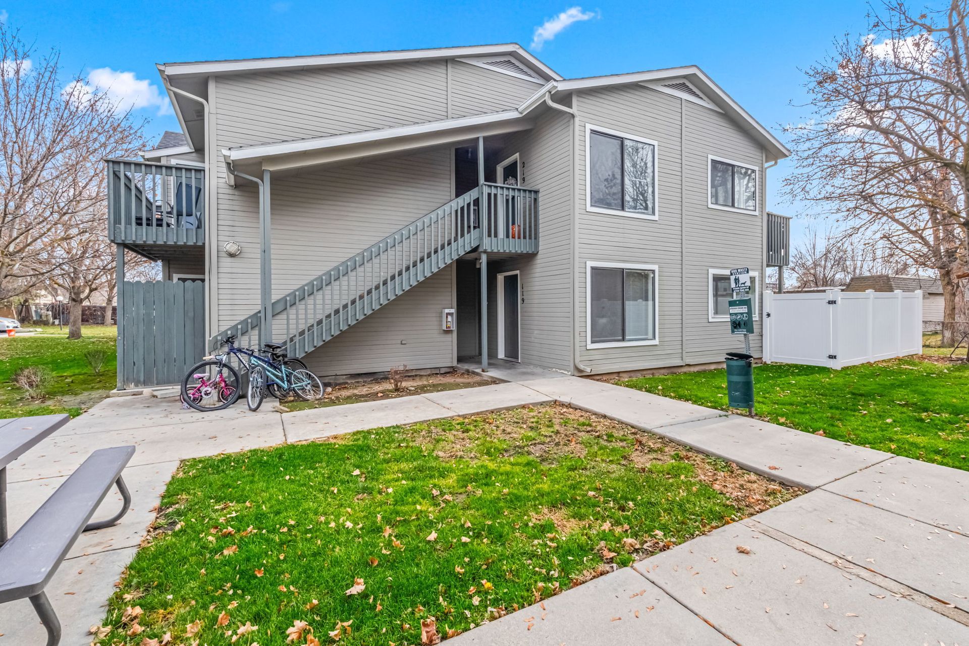 Two-story apartment building with stairs, bikes, and a grassy area in front.