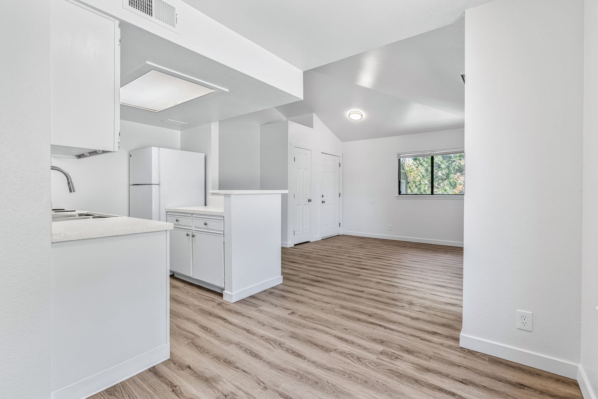 Interior view of a white kitchen with a wooden floor and a view of a living space with a window.
