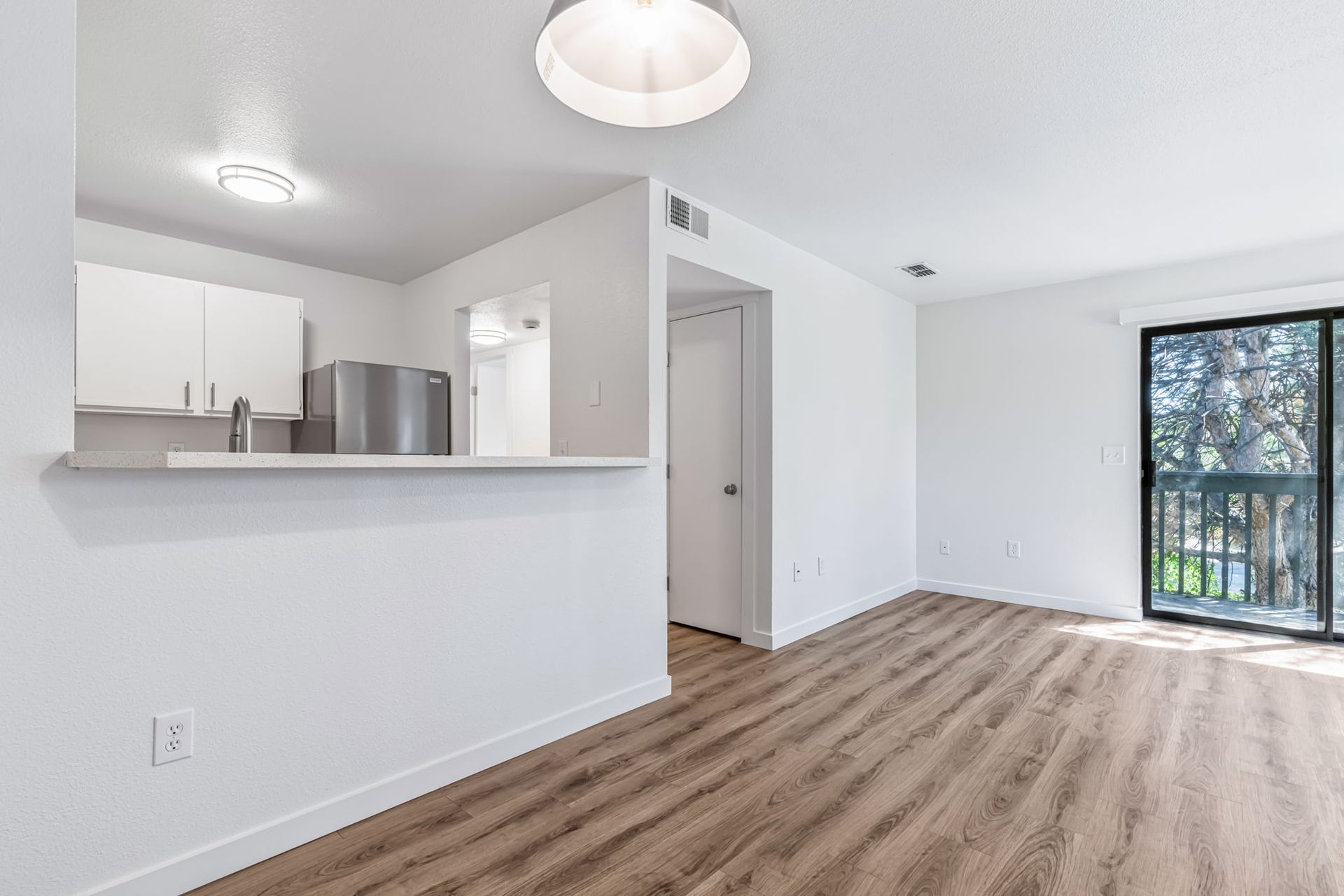 Bright, empty apartment interior with wood-look flooring, white walls, and a view of trees from a sliding glass door.