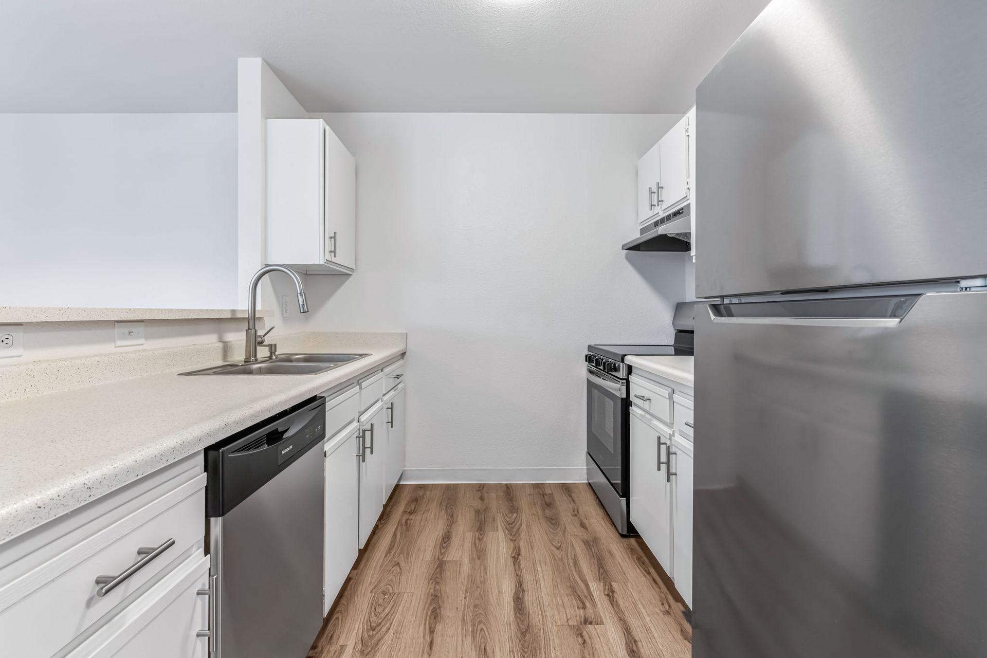 A narrow, white kitchen with stainless steel appliances, light-colored countertops, and wood-look flooring.