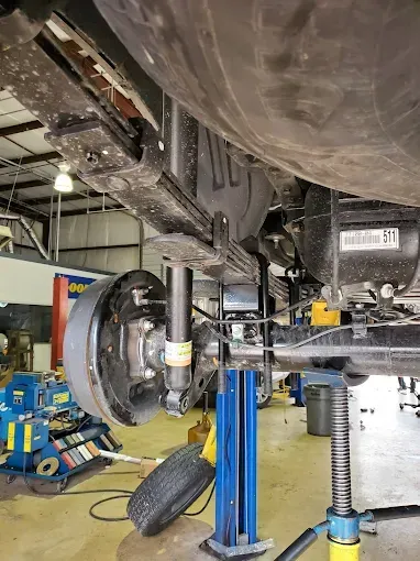 Underside of a vehicle in a garage. Rear suspension, axle, and tire visible. Supported by a blue jack.
