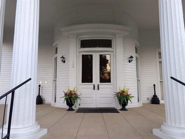 The front door of a white house with columns and potted plants in front of it.