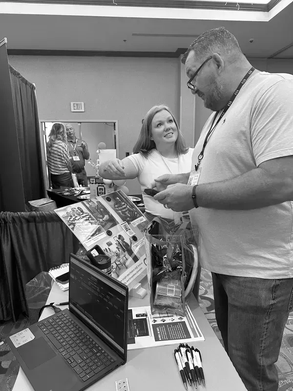 Two people chatting at a booth with a laptop, brochures, and product displays at an indoor event.
