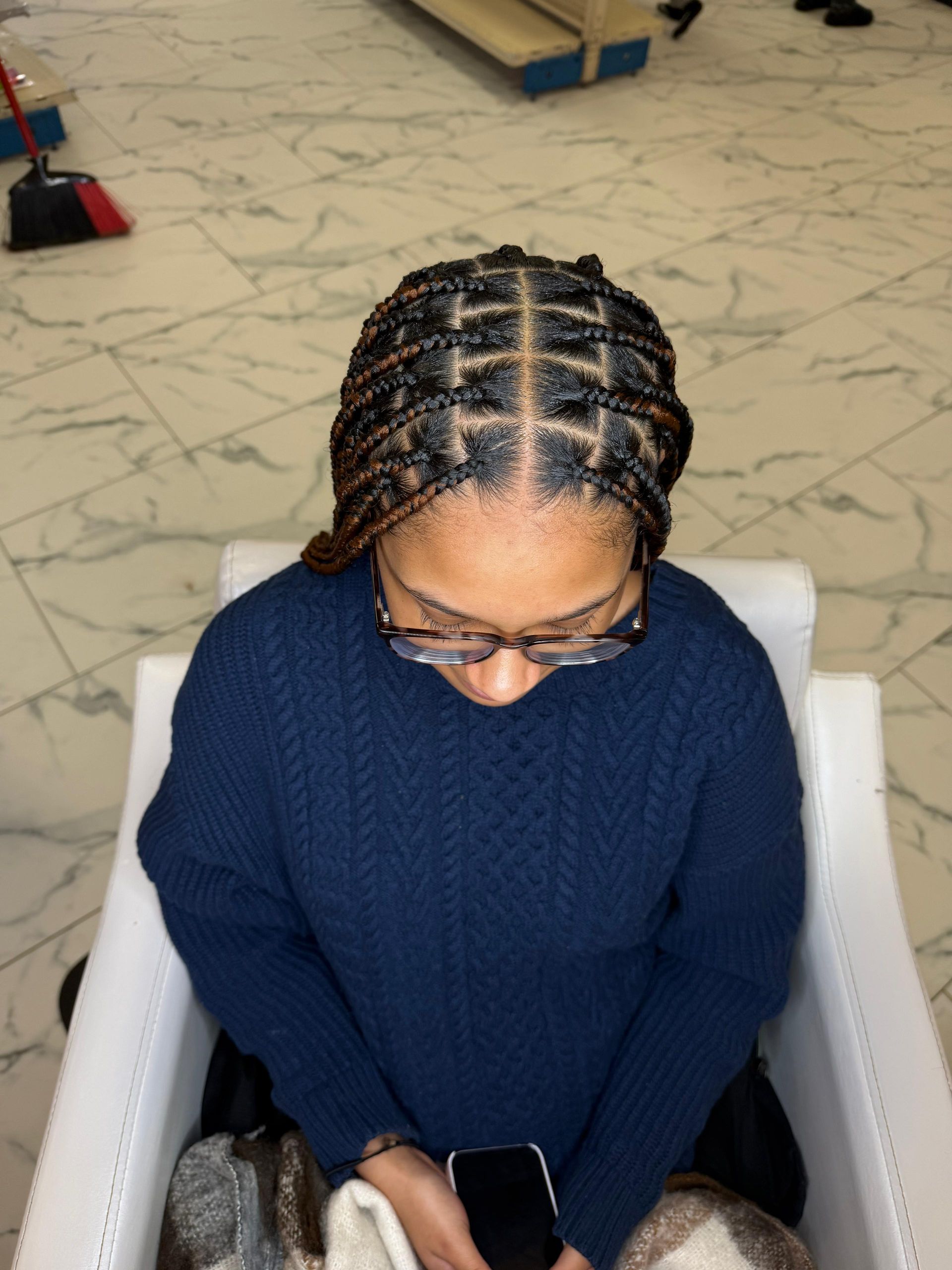 Person with braided hair wearing glasses and blue sweater seated in white chair.