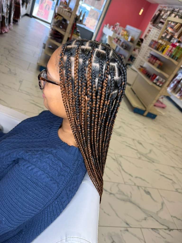 Woman with box braids in a hair salon. Braids are brown and black. Wearing glasses and blue sweater.
