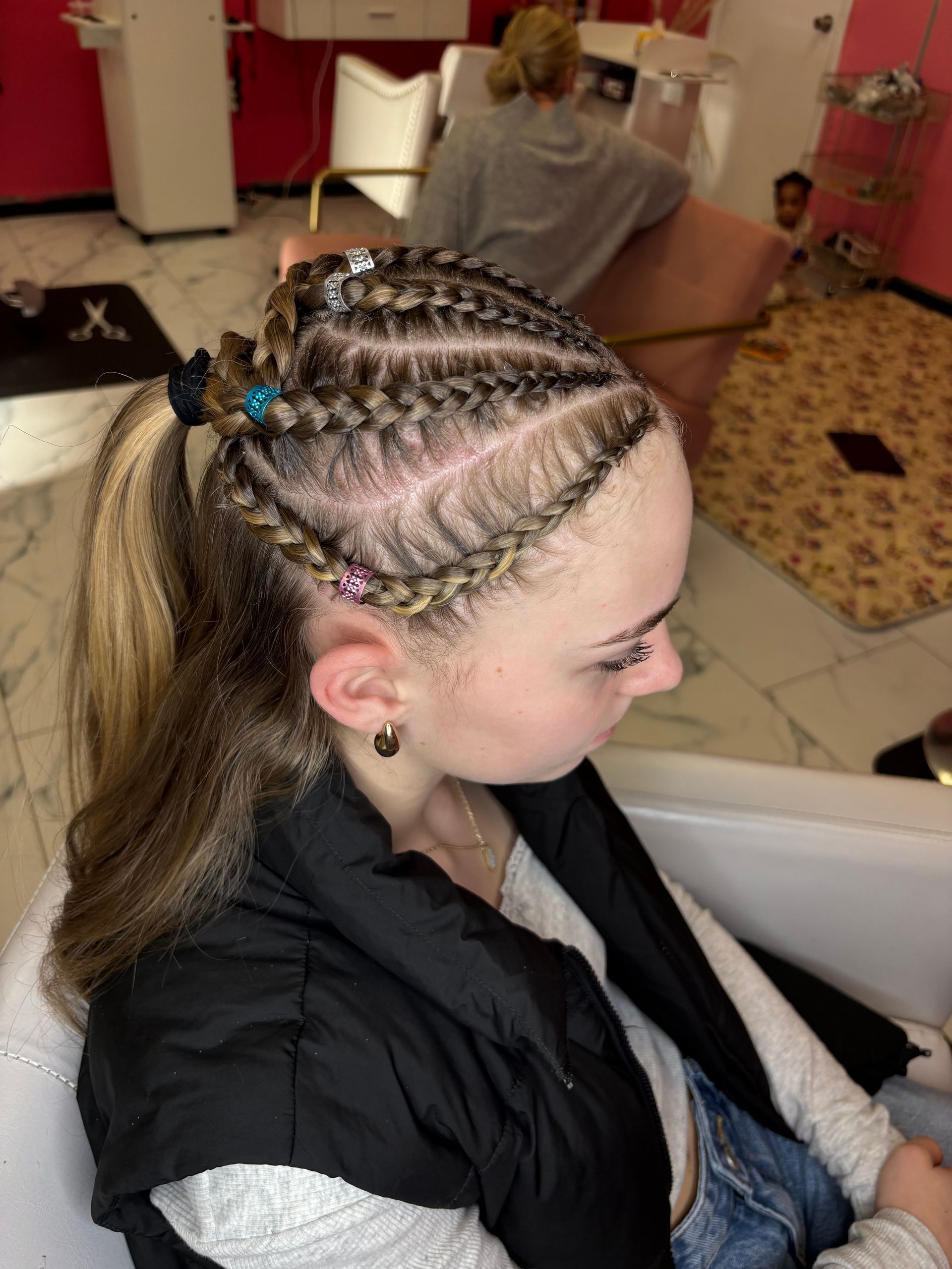 Woman with braided hairstyle, sitting in a salon.  Braids with beads, wearing a black vest and light blue jeans.