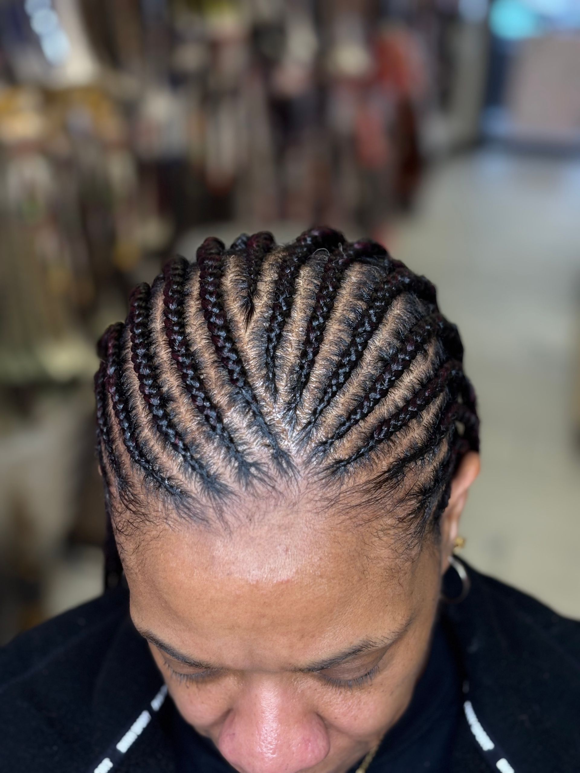 Close-up of cornrow braids on a person's head, angled from above. Braids are dark. Setting appears to be indoors.