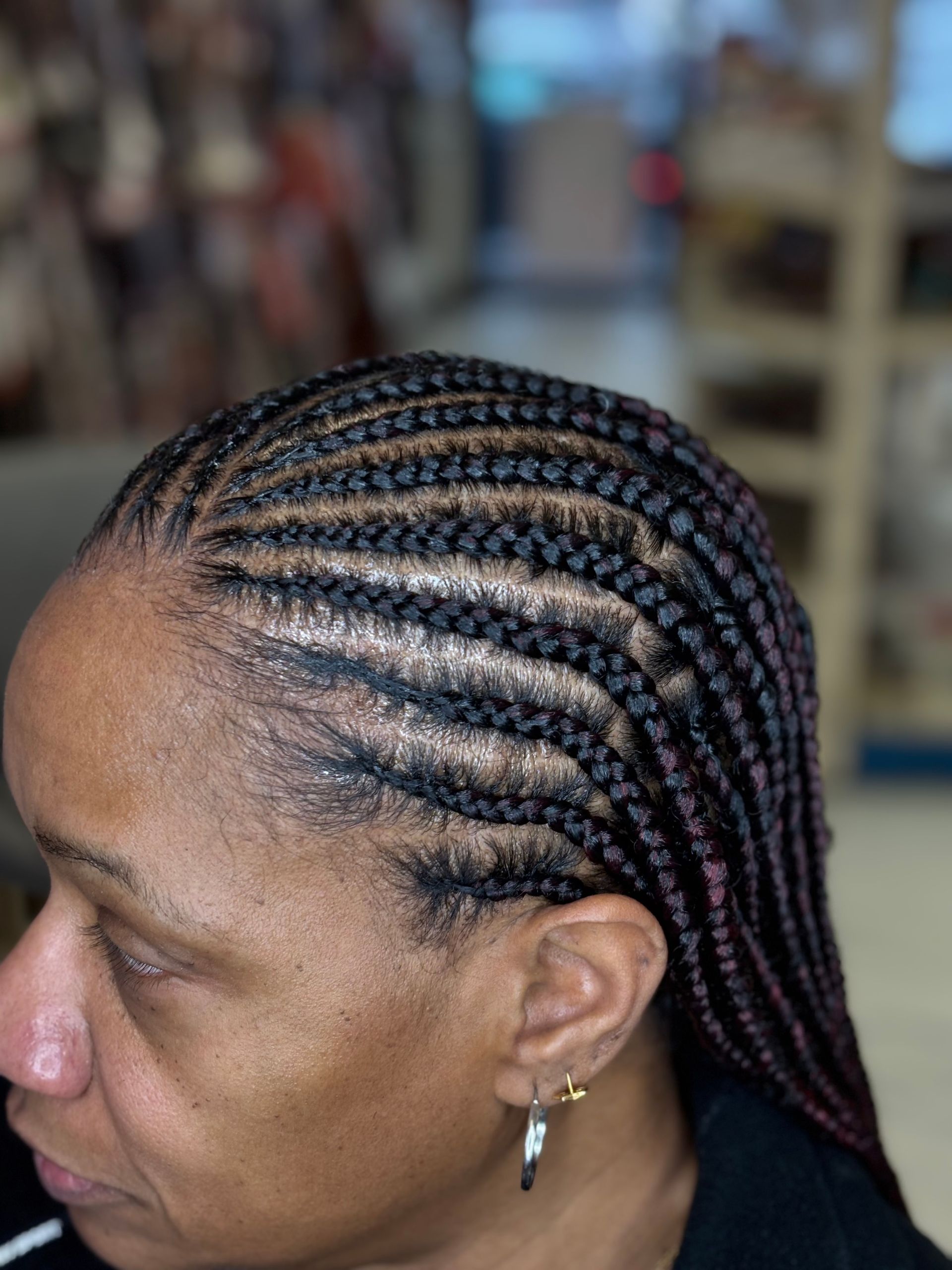 Braided hairstyle on a person's head, close-up. Rows of braids. Tan skin, visible ear. Indoors, blurred background.