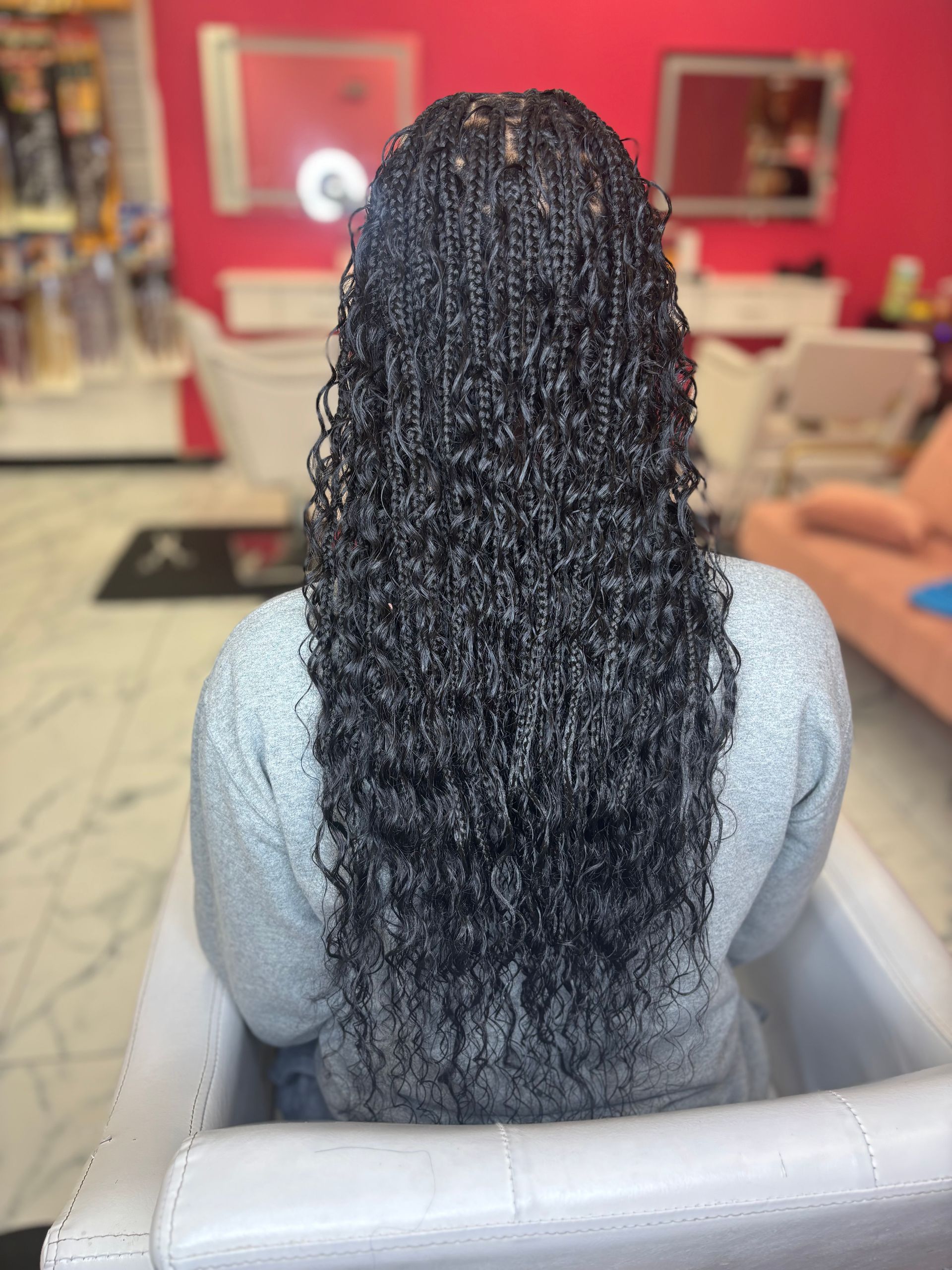 Back view of a person with long, black micro braids with curly ends, seated in a salon chair.