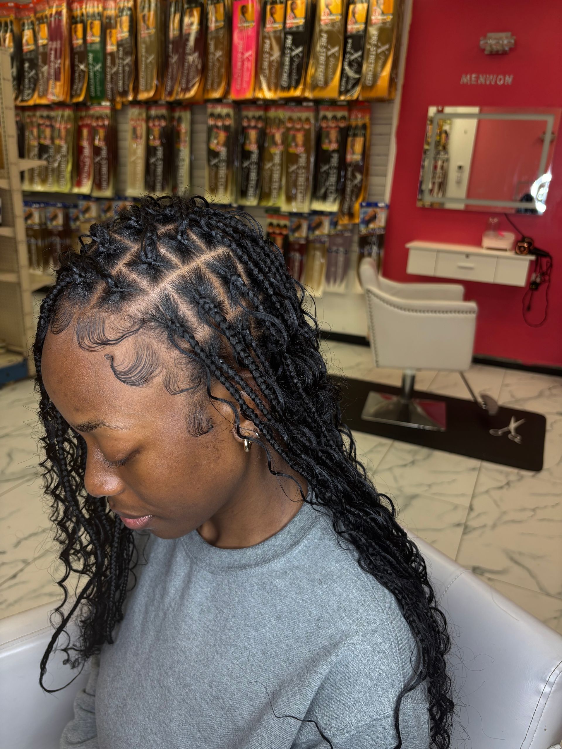 Woman with small, curly black braids in salon, posing with head down, some decorative braid patterns on top.