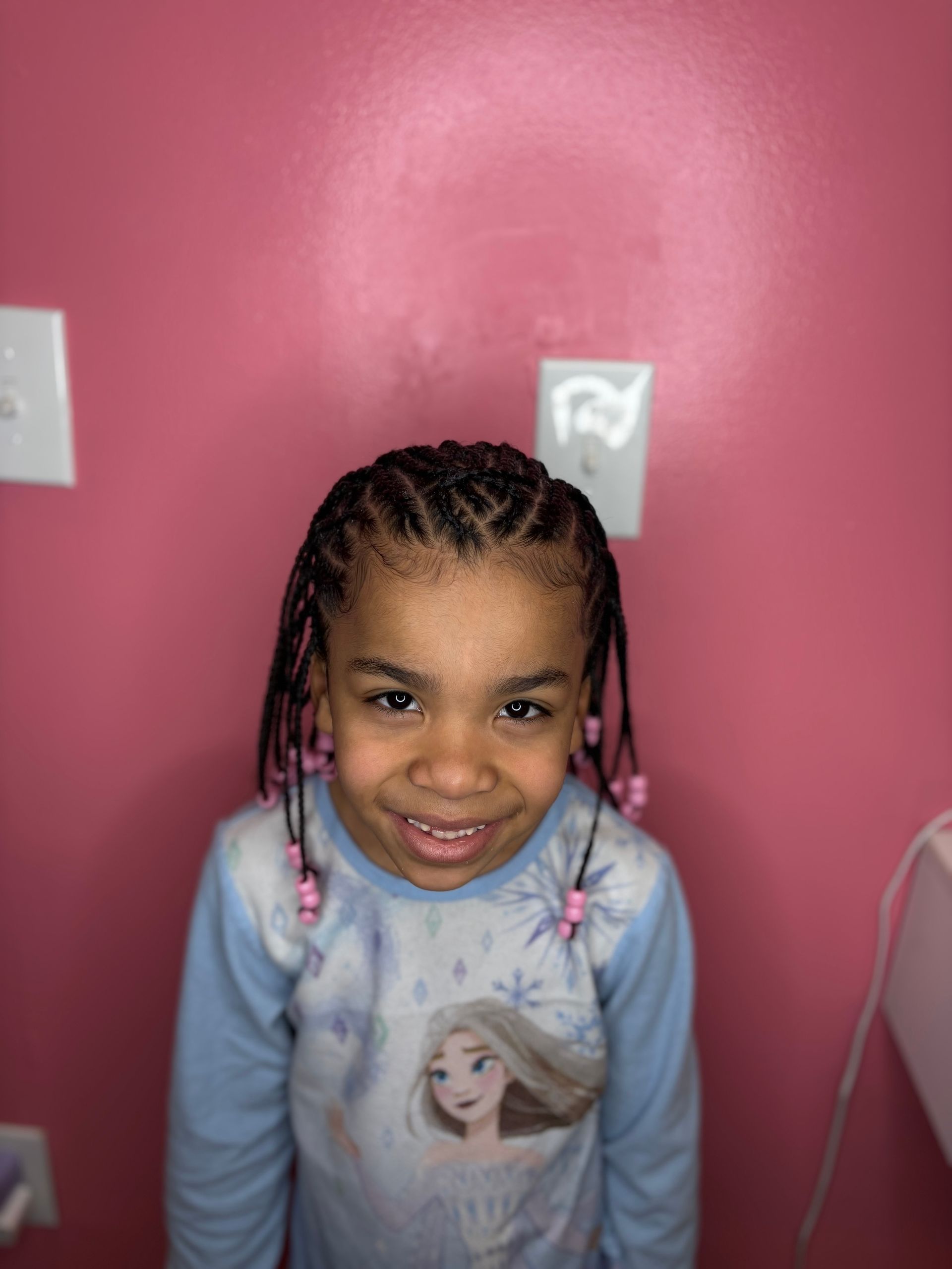 Girl with braided hair and pink beads smiles in front of a pink wall, wearing a light blue Frozen shirt.