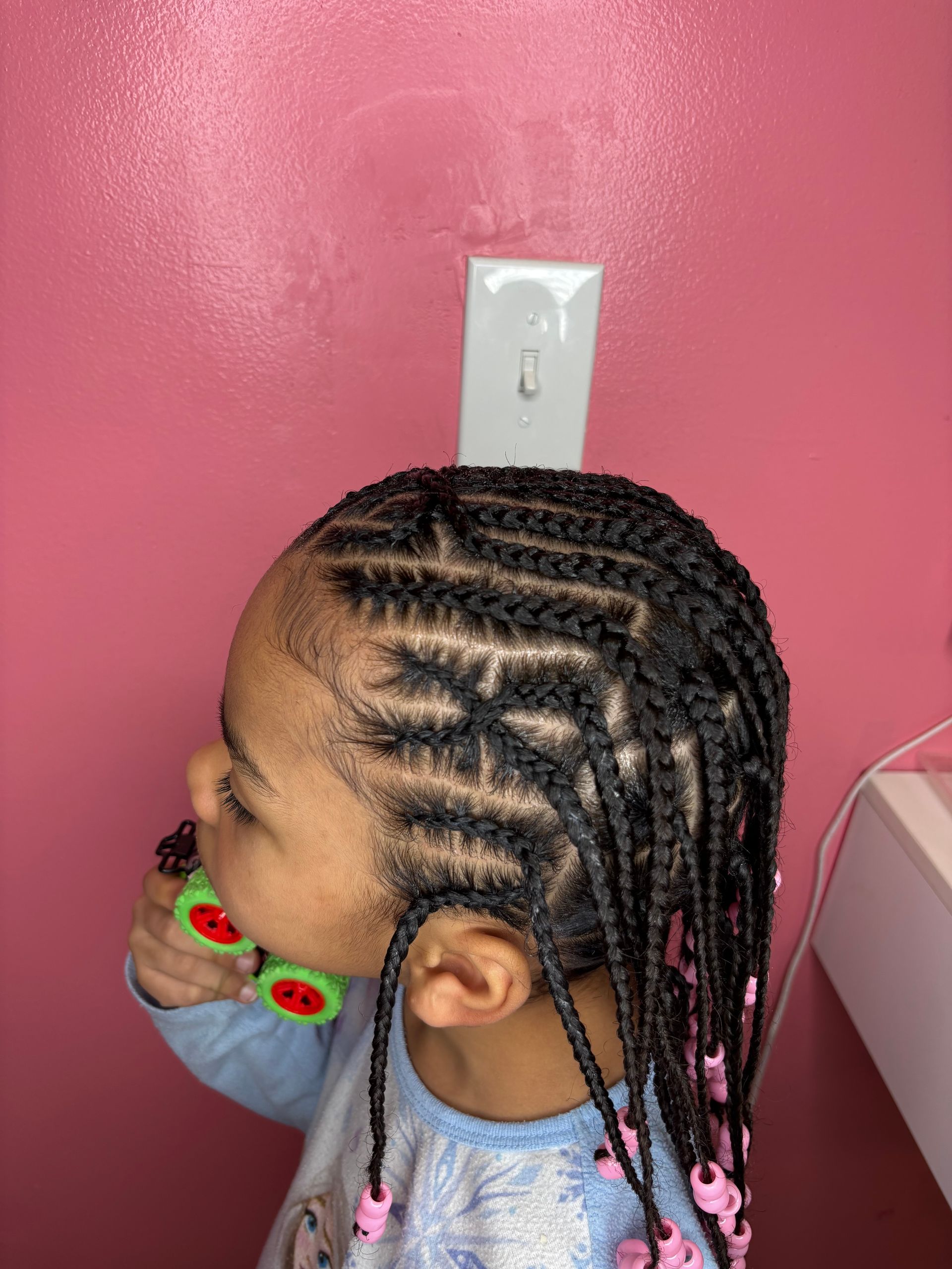 Girl with braided hair, biting toy in front of pink wall.