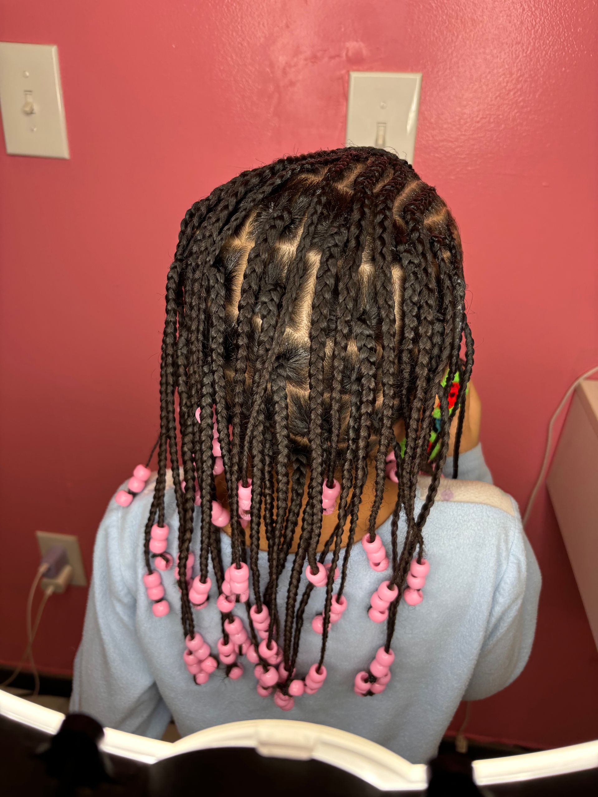 Braided hairstyle with pink beads. Child's back is to the camera against a pink wall.