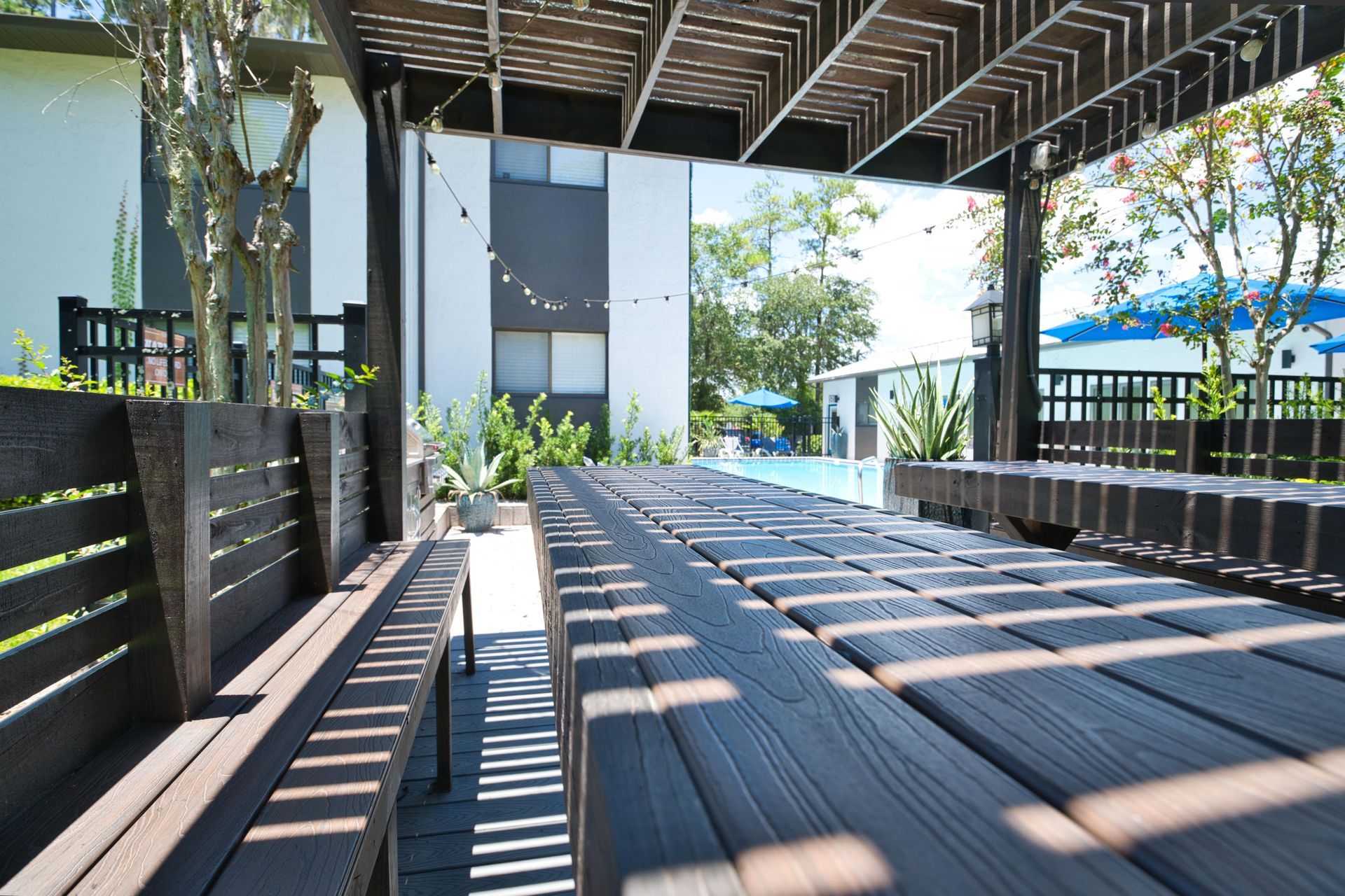 A long wooden table under a pergola with a pool in the background