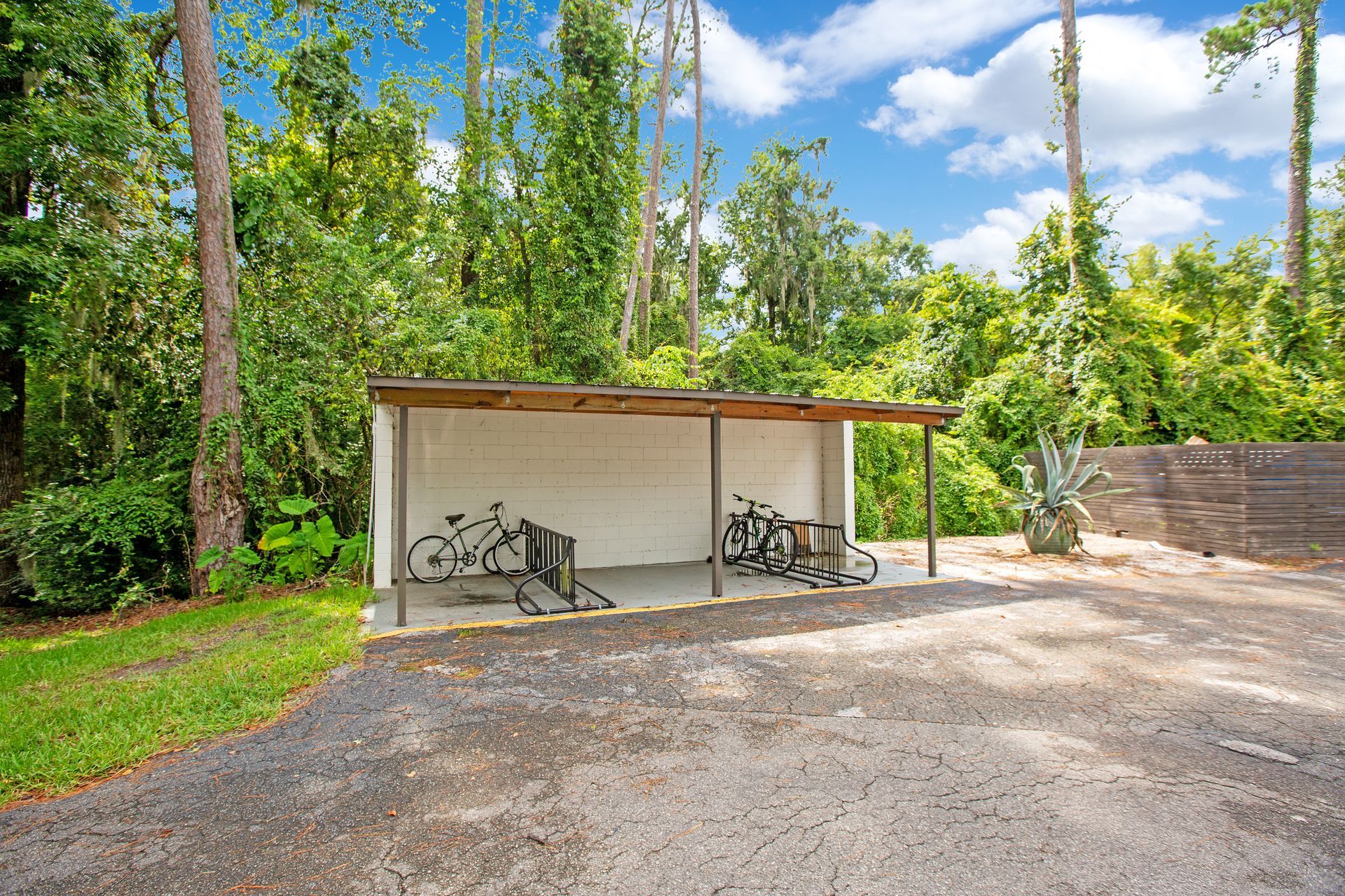 A shed with bikes parked underneath it in a parking lot surrounded by trees.