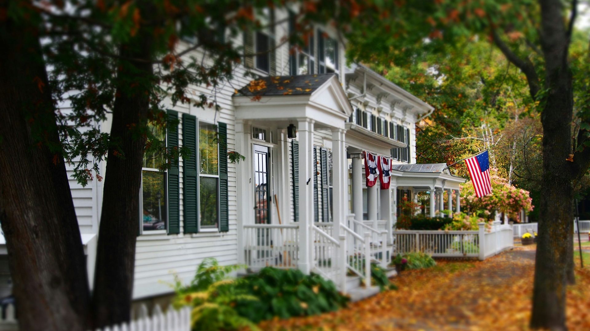 A white house with green shutters and an american flag on the porch.