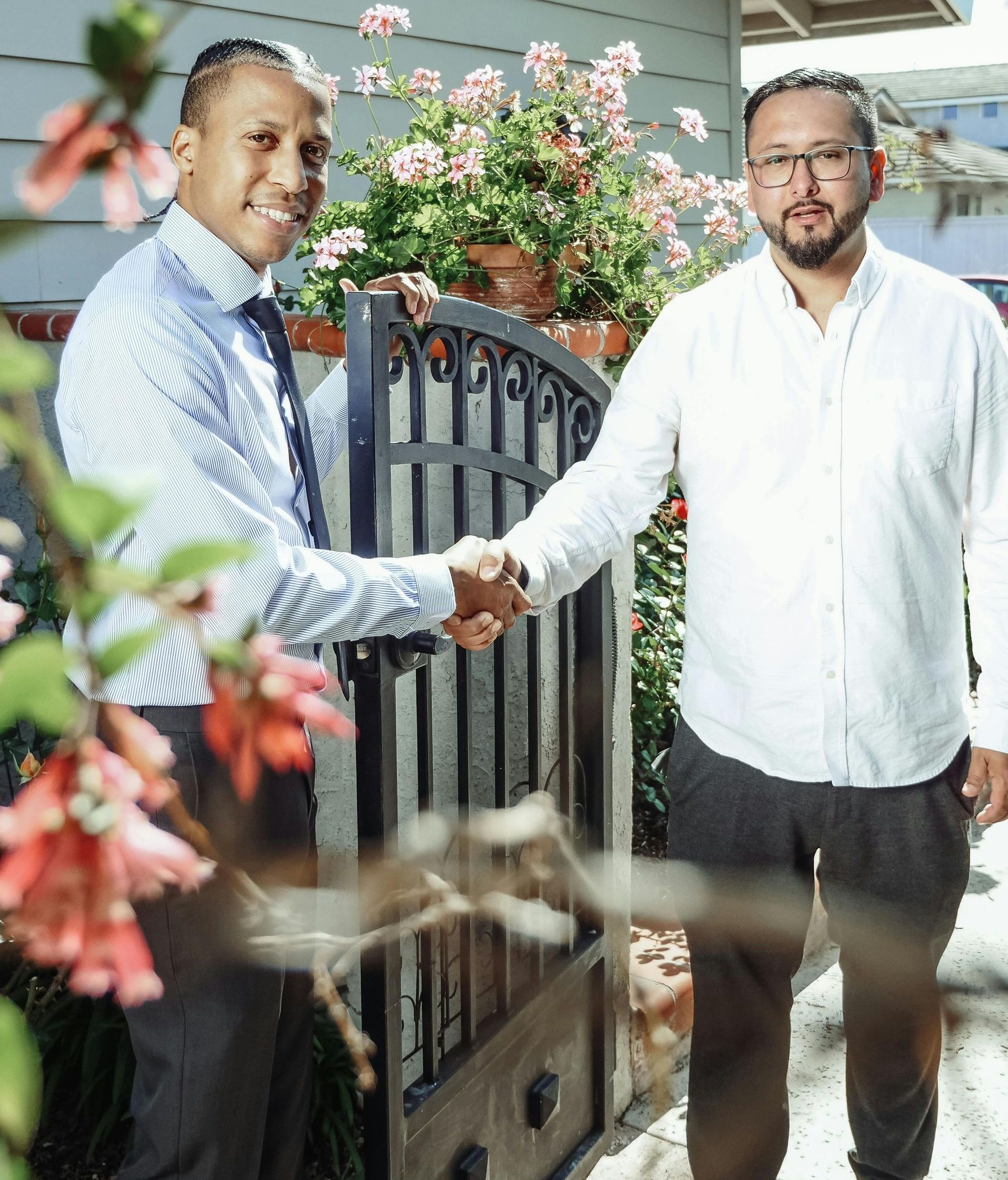 Two men shaking hands in front of a gate with flowers in the background