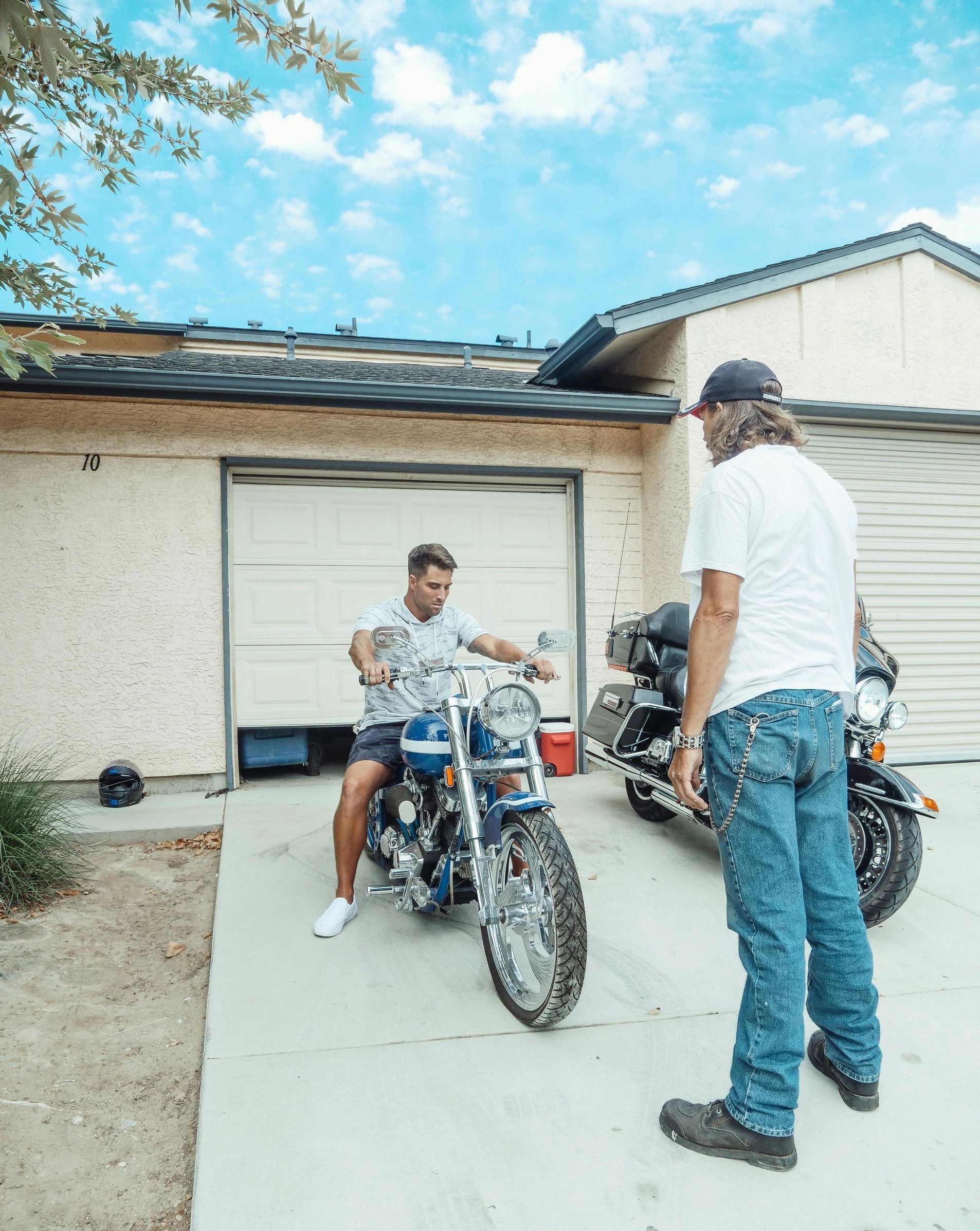 A man is standing next to a man on a motorcycle in front of a house.