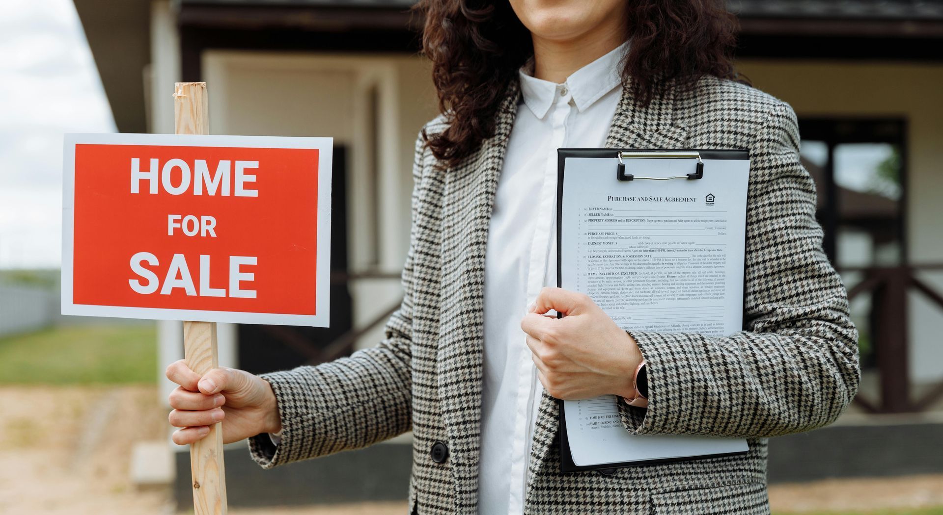 A woman is holding a home for sale sign and a clipboard in front of a house.