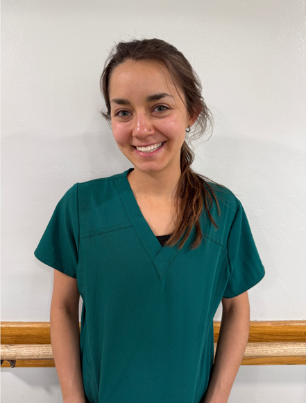 Smiling person in navy scrubs with a badge, standing against a dotted beige wall.