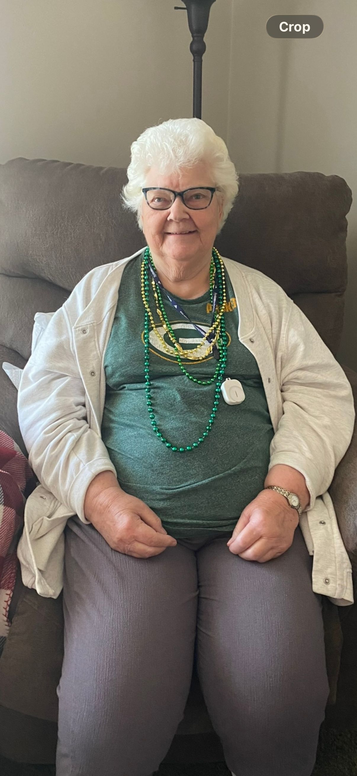 Smiling person seated on a couch, wearing glasses, a white cardigan, and green top with necklaces.