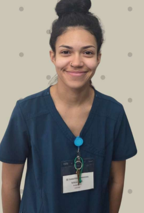 Smiling person in navy scrubs with a badge, standing against a dotted beige wall.