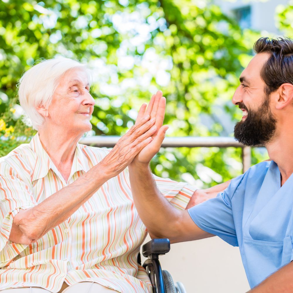 An elderly woman in a wheelchair is giving a high five to a nurse.