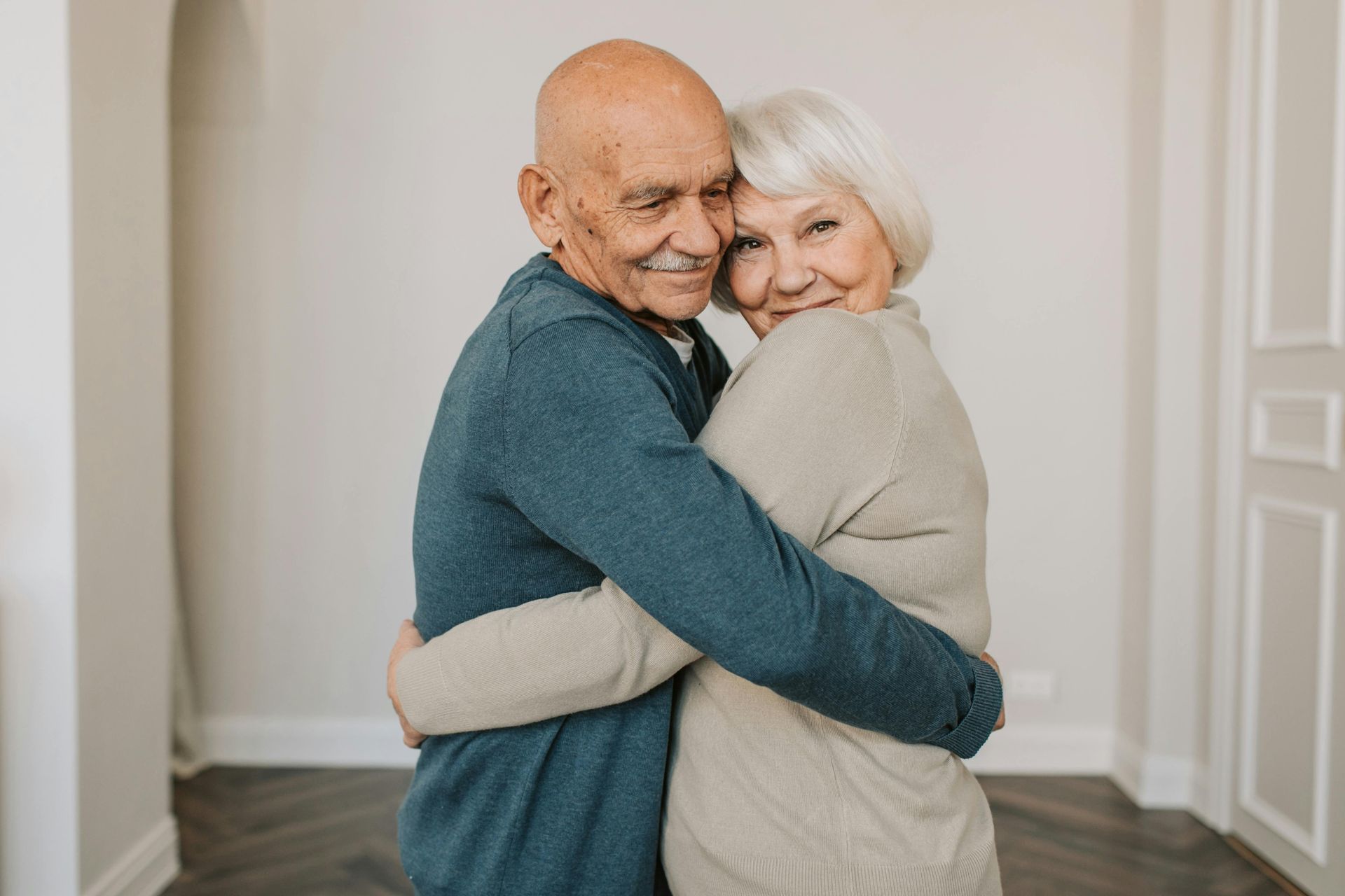 An elderly couple is hugging each other in a living room.