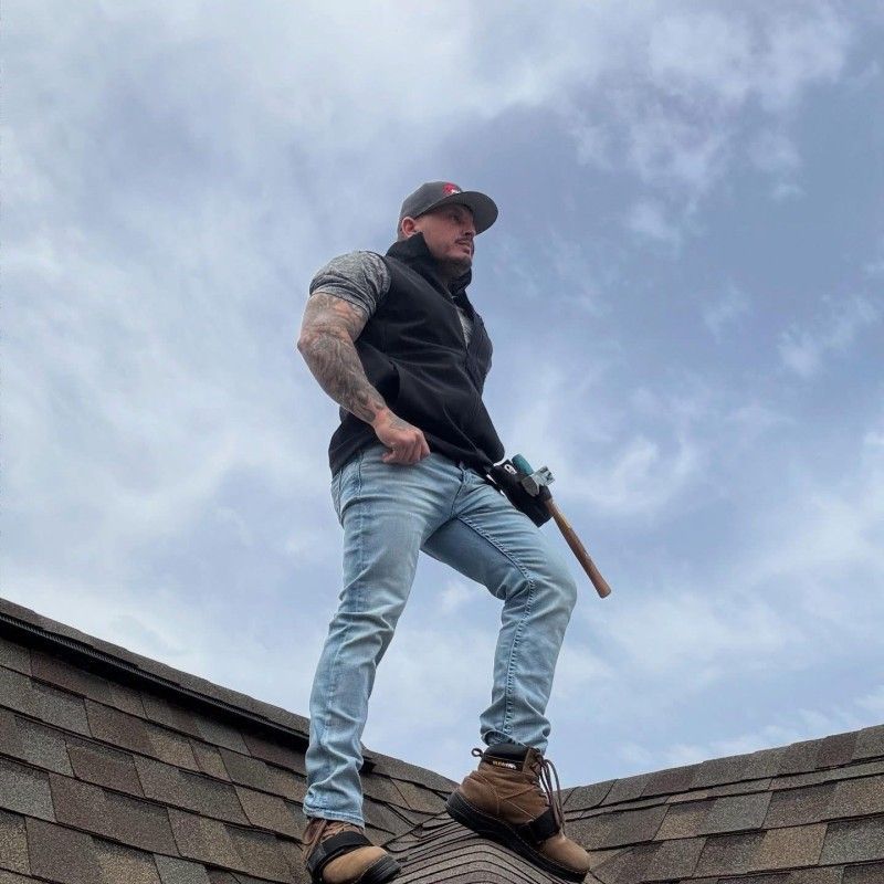 Man on a roof, holding a hammer, wearing a vest, jeans, and a cap, with cloudy sky background.