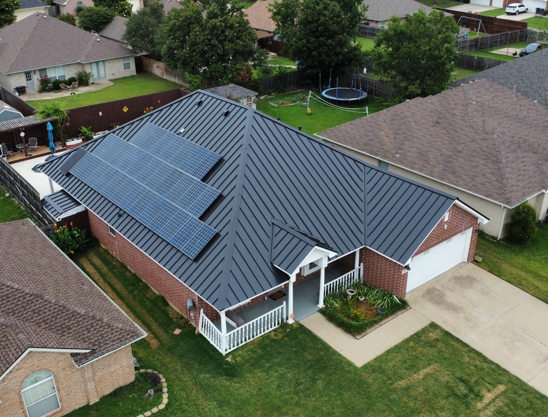House with solar panels on a dark gray metal roof. Brick exterior, front yard, suburban setting.
