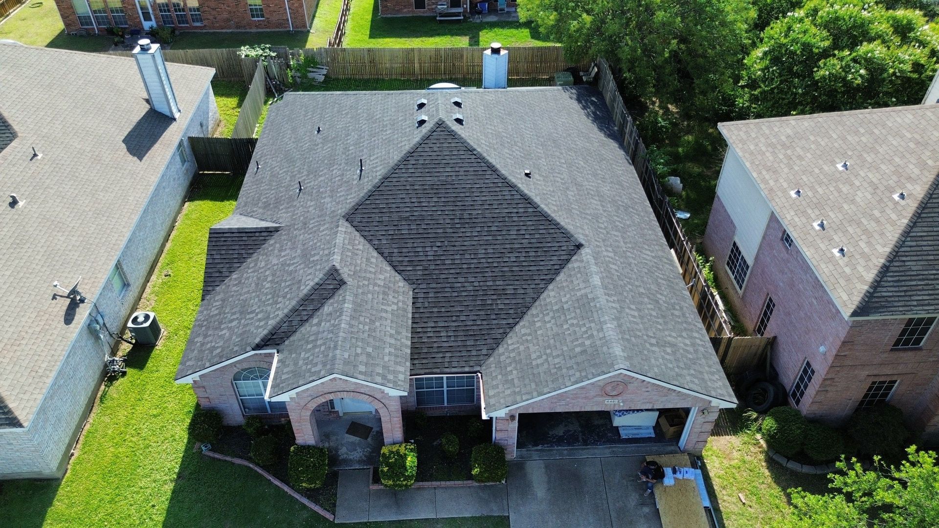 Aerial view of a residential home with a dark roof, surrounded by trees and other houses in a suburban setting.