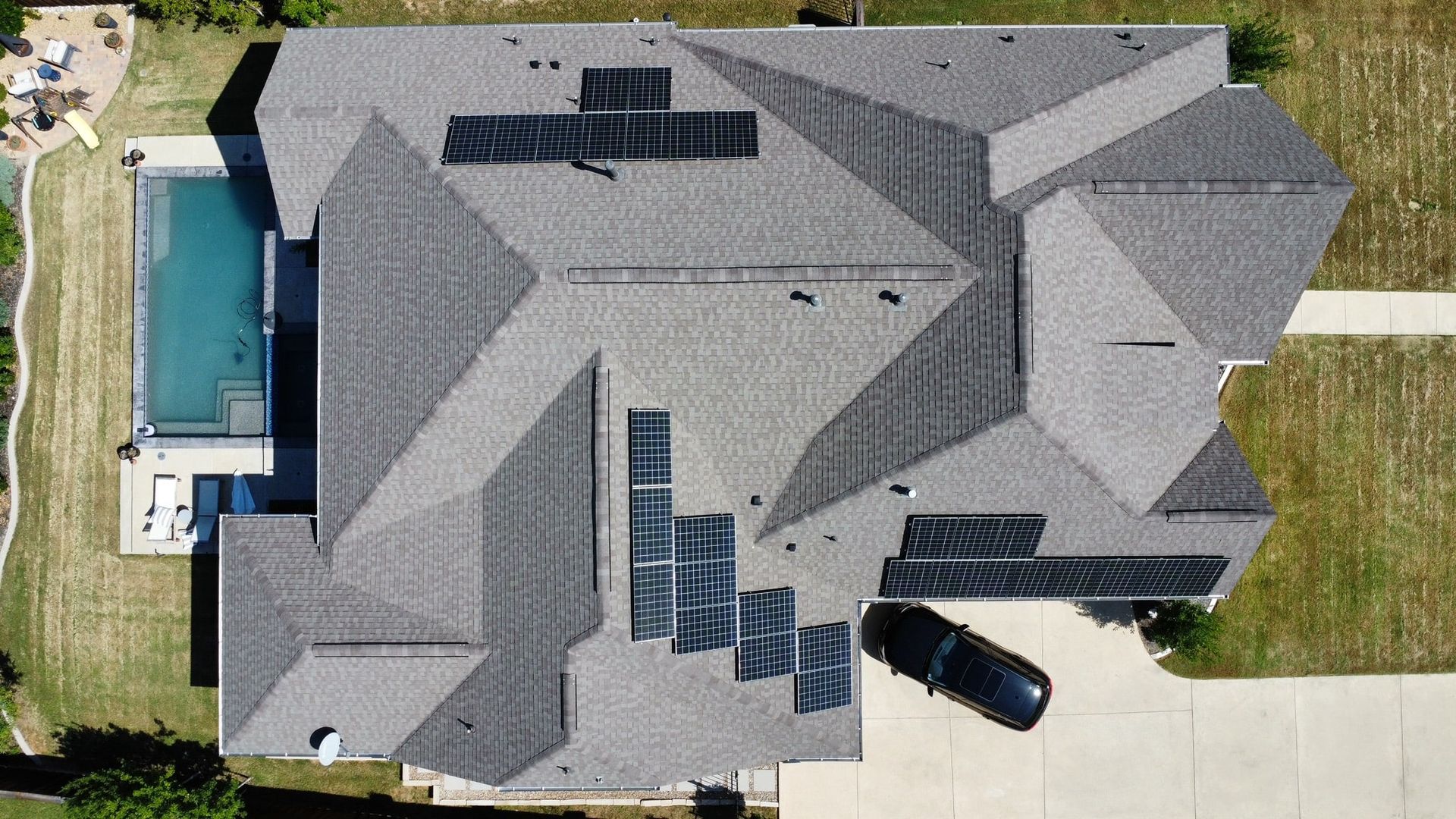 Aerial view of a gray-roofed house with solar panels and a pool, black car in the driveway.